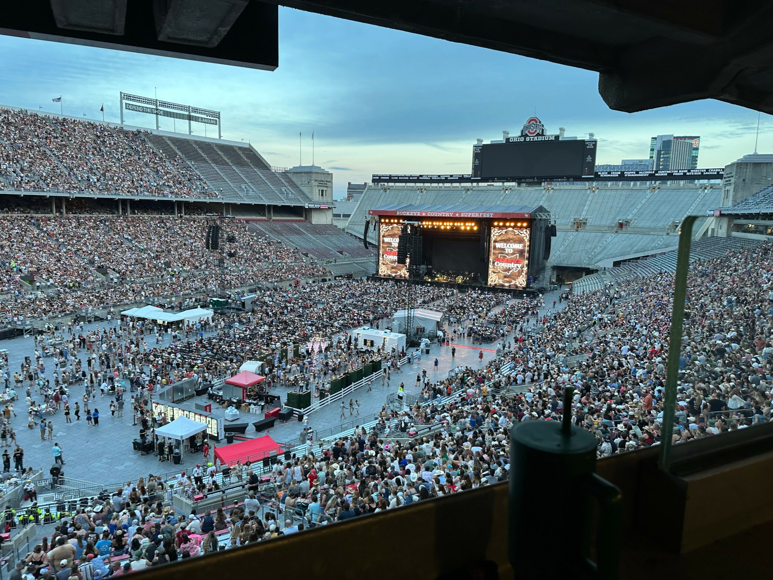 Large crowd gathered in a stadium for a concert with a stage and large screens, viewed from a balcony.
