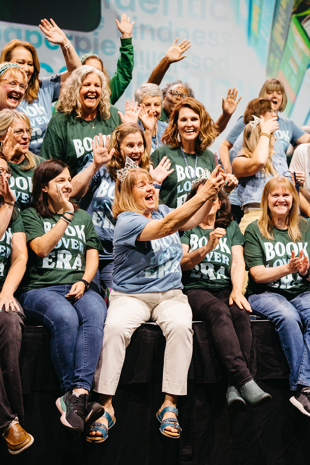 Group of women and girls smiling, cheering, and raising hands on stage, wearing matching green and black shirts, celebrating.