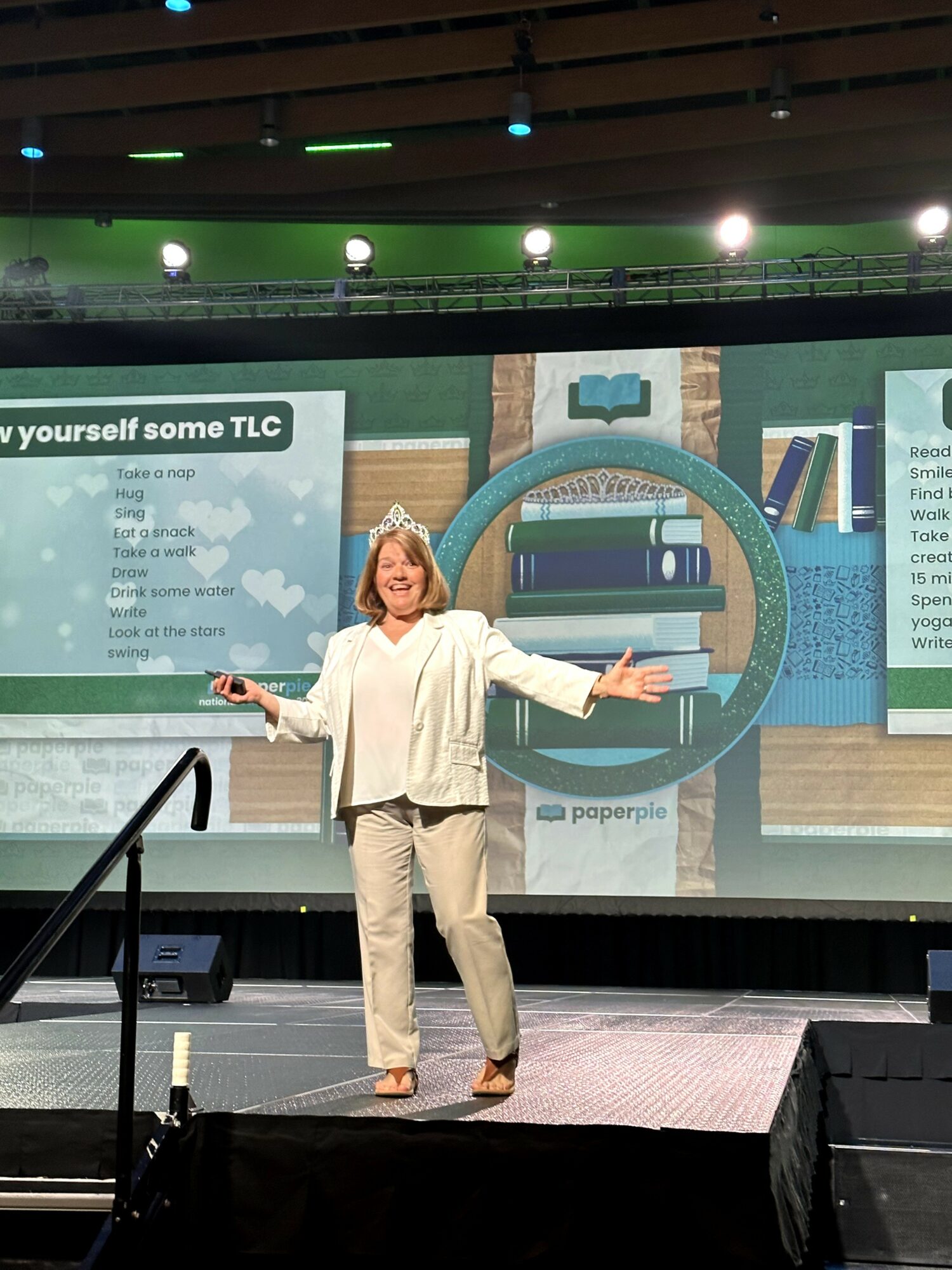 Woman standing on stage with arms outstretched, background features a large screen with books and text, stage lighting overhead.