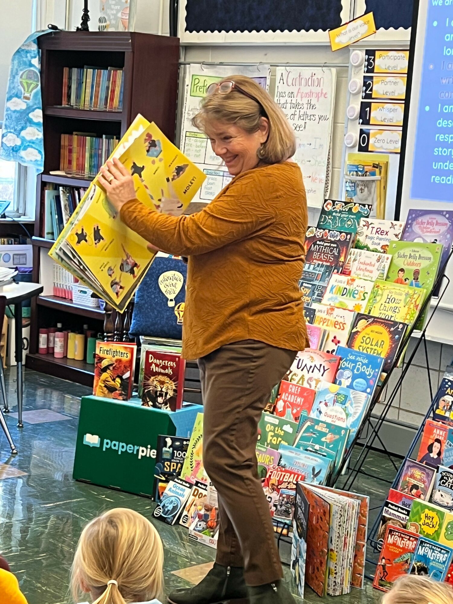 Woman reading a children's book in a bookstore with shelves of books around her, a child nearby, and a green sign.