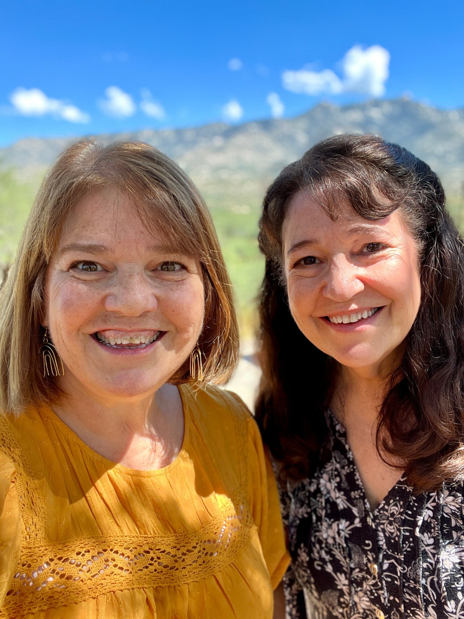 Two women smiling outdoors with mountains and blue sky in background.
