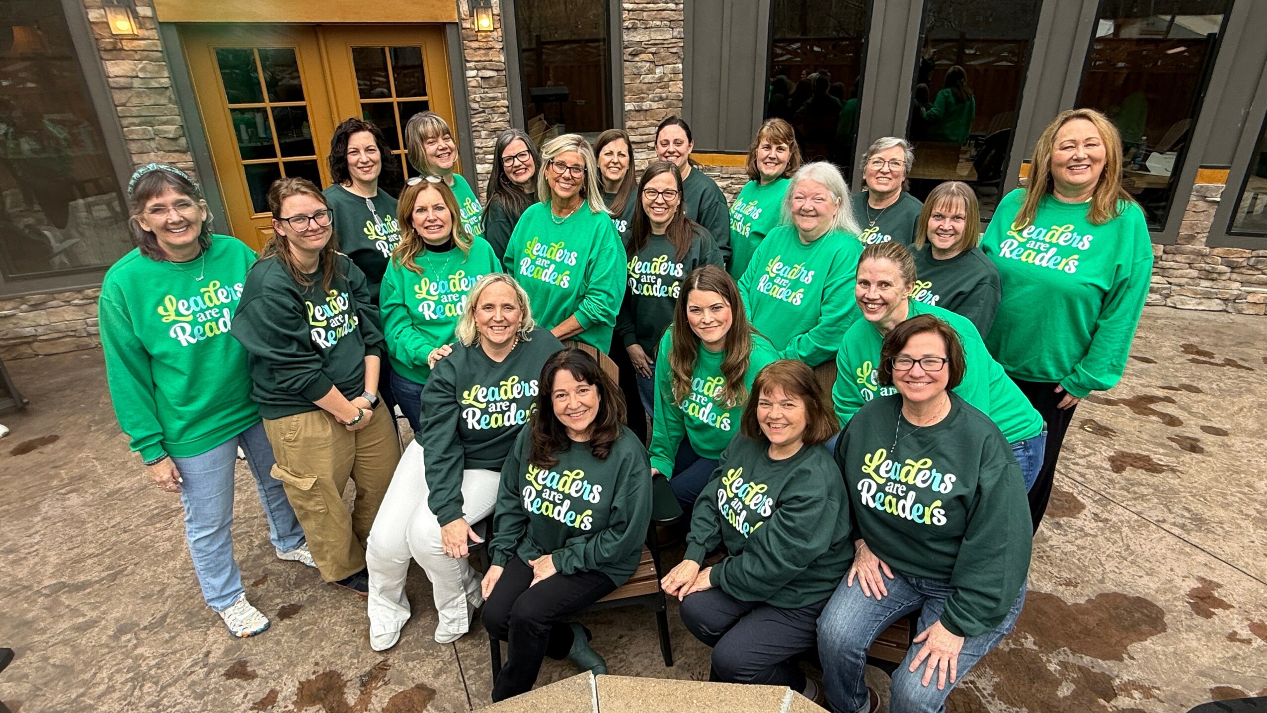 Group of 20 people outdoors, some wearing green and black sweatshirts, smiling and posing for a photo.