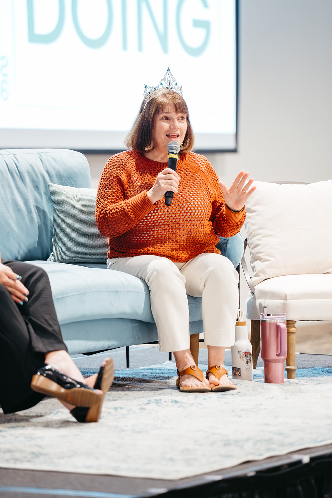 Young girl wearing a crown and orange sweater, speaking into a microphone while sitting on a light blue sofa.