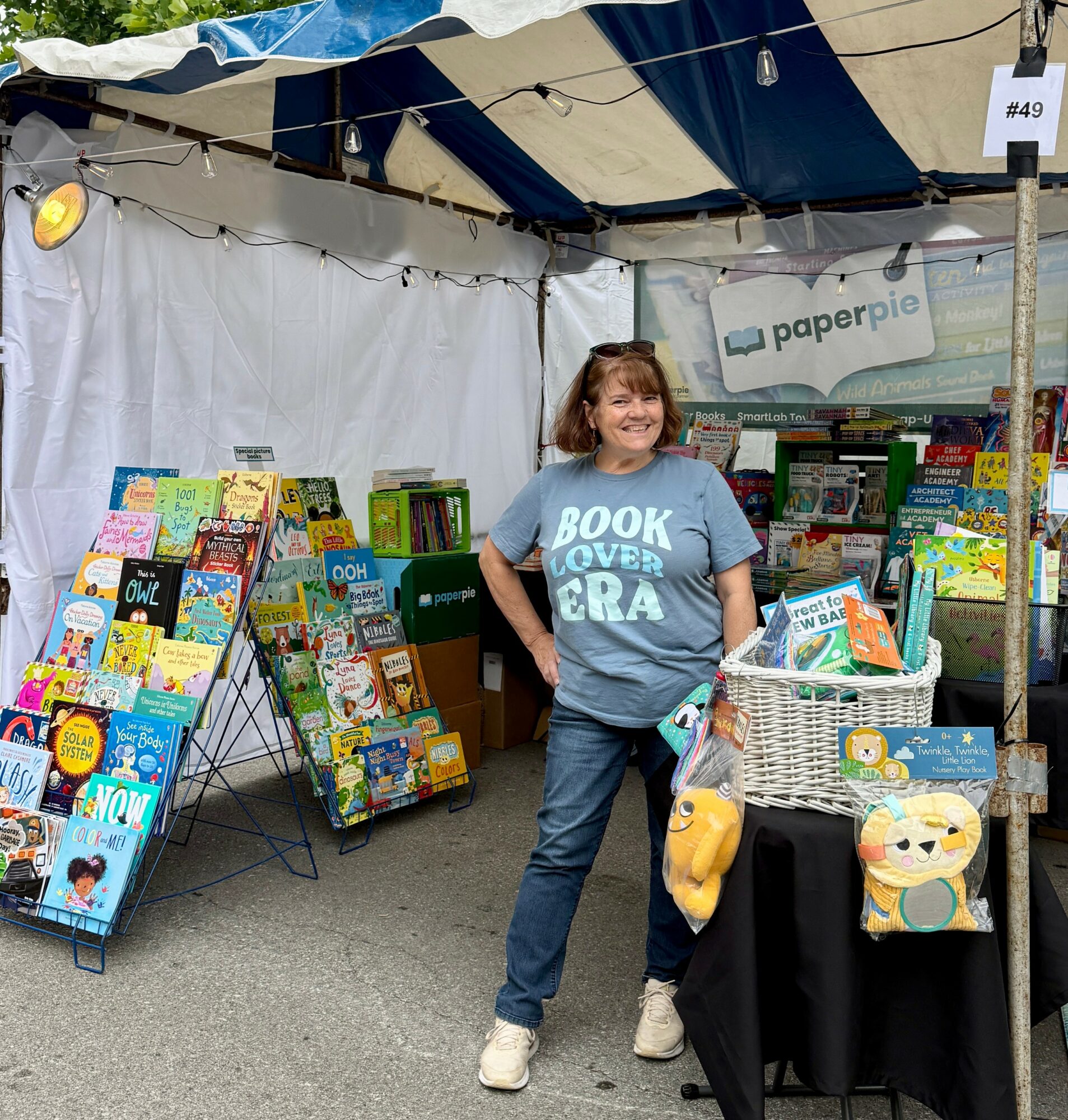 Woman standing at a book stall outdoors, smiling, with books displayed on racks and a table, under a canopy.