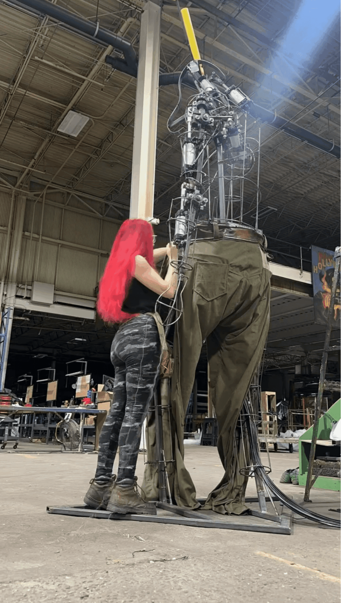 Person with long red hair working on a large mechanical structure in an industrial setting.