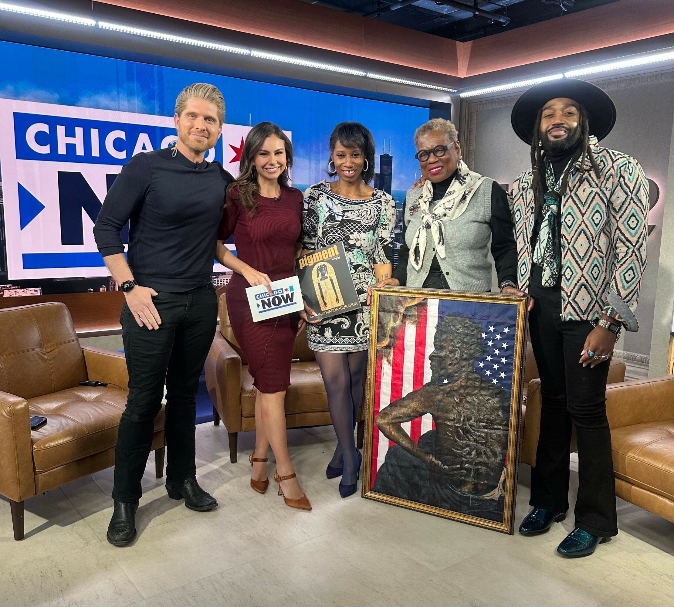 Five people stand in a TV studio with a framed artwork of a woman in front of an American flag background, smiling.