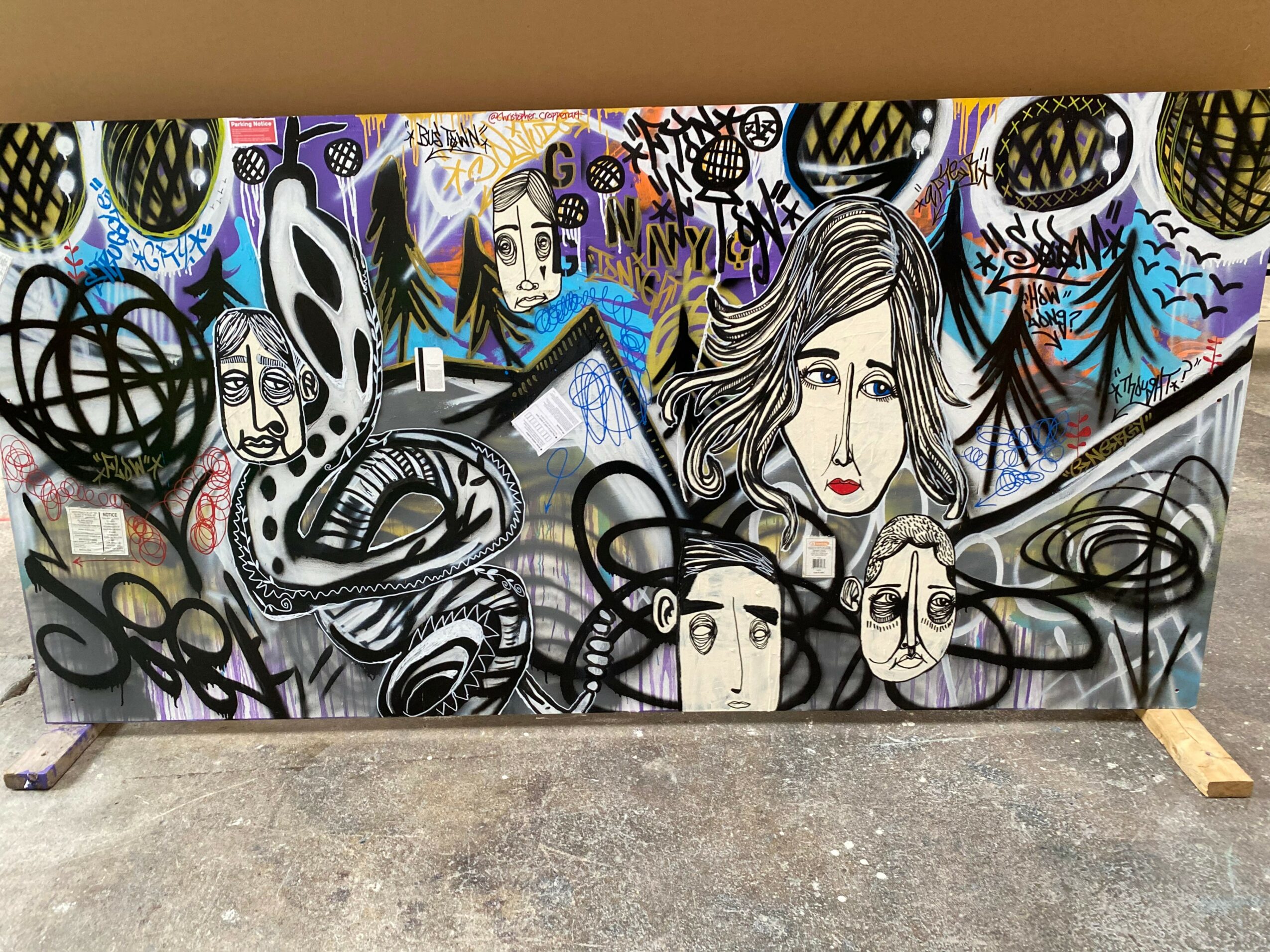 Colorful graffiti mural with faces, abstract shapes, and patterns on a wall, with a concrete ground below. Alt text word count: 20.