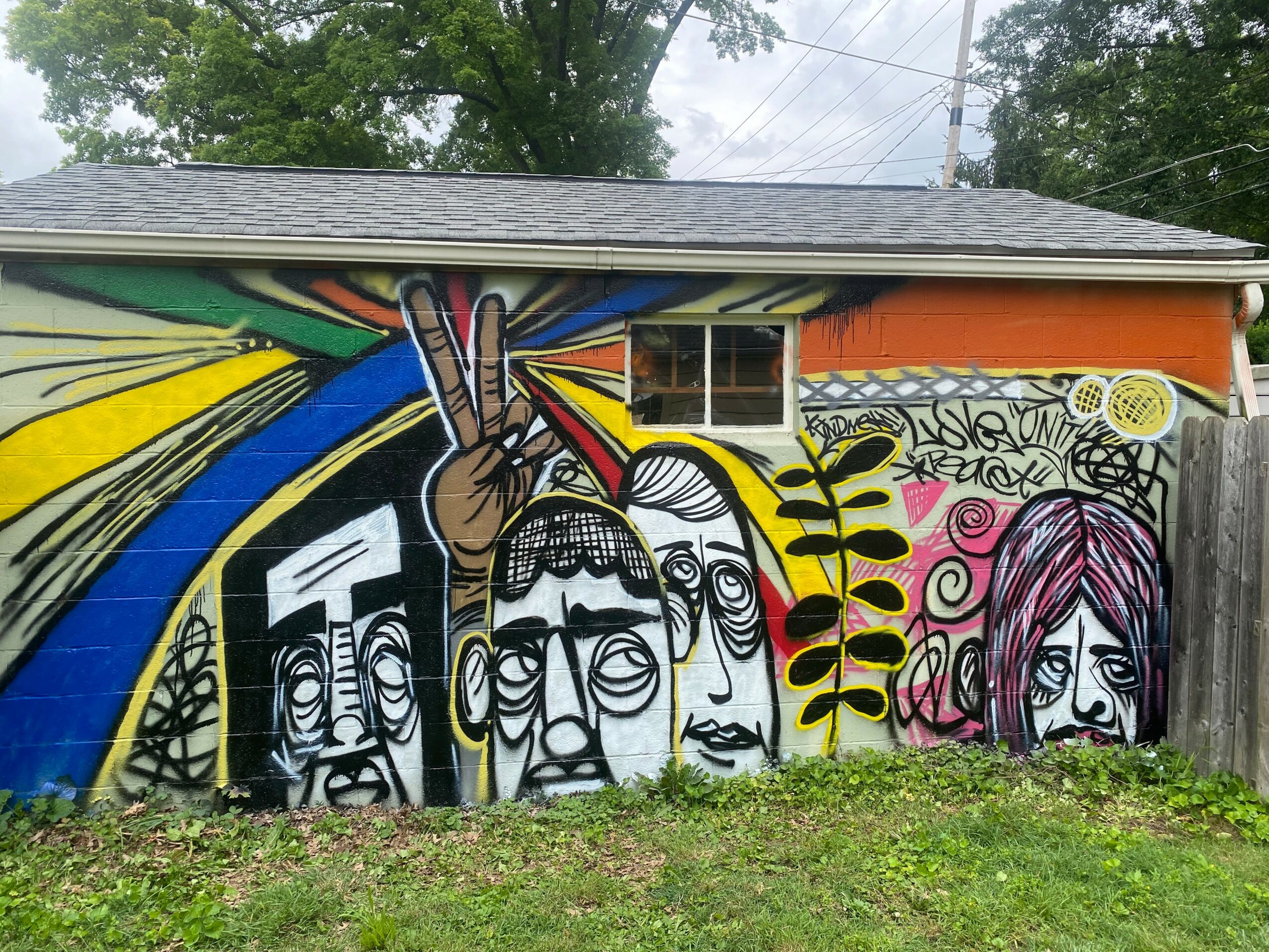 Colorful mural of stylized faces and a hand making a peace sign on a building wall, with trees and sky in background.