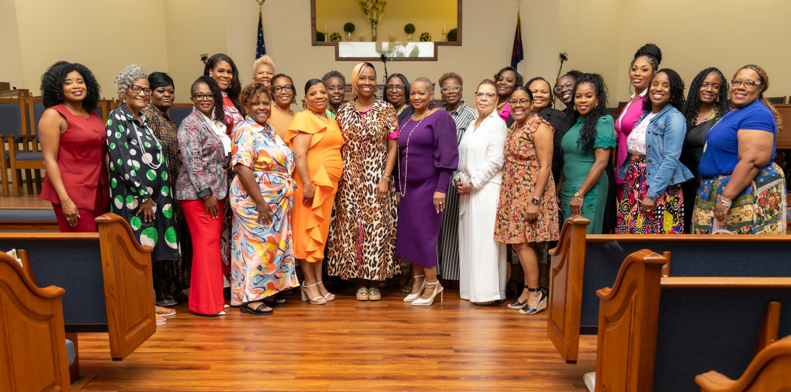 Group of women standing in a room with wooden floors and flags, smiling for a photo.