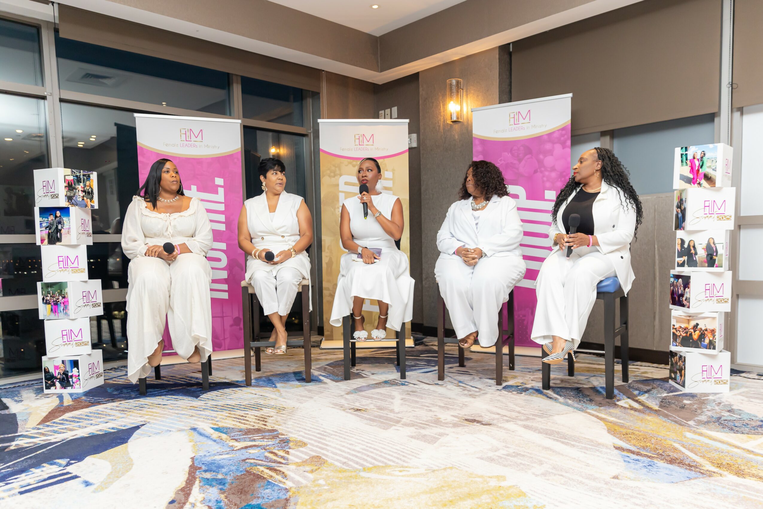 Five women sit on chairs in a panel discussion, with pink and white banners behind them, in a room with a patterned carpet.