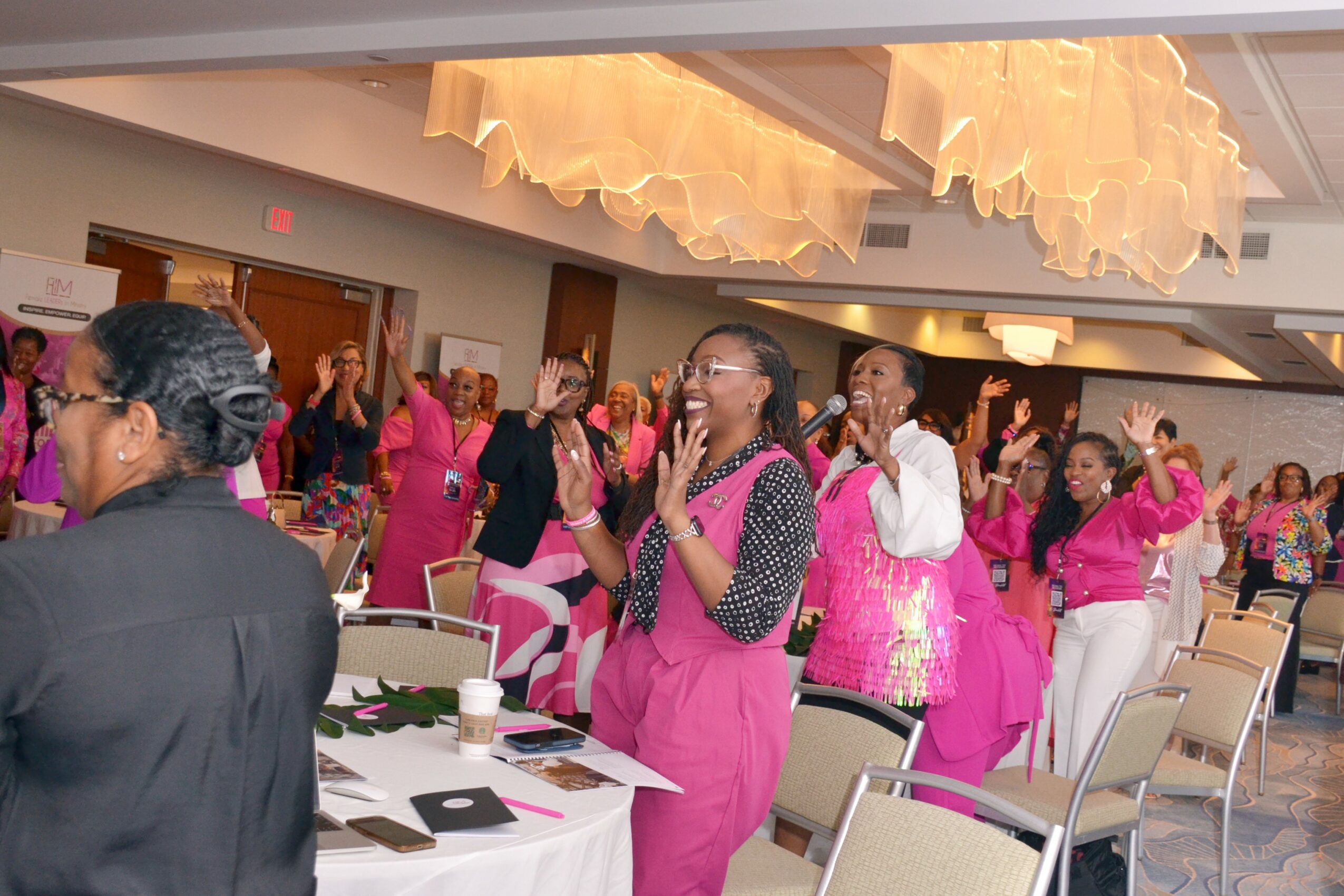 Group of women wearing pink aprons and white clothing, dancing and celebrating in a decorated indoor event space.