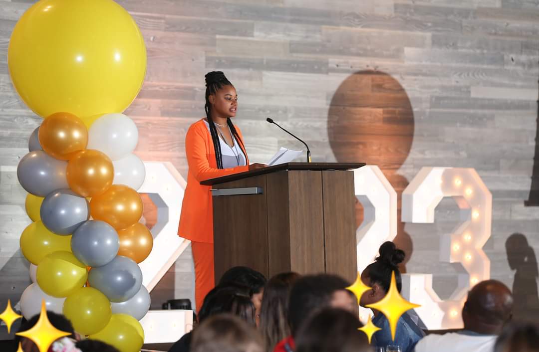 Woman speaking at podium during event with large balloon decoration and audience, background with wooden wall and large illuminated numbers.