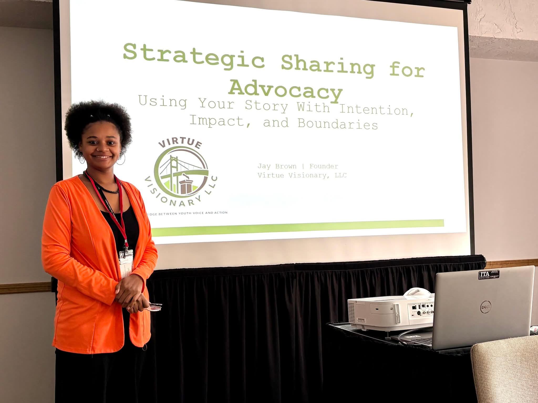 Woman standing in front of a presentation screen with a slide titled 'Strategic Sharing for Advocacy' in a conference room.