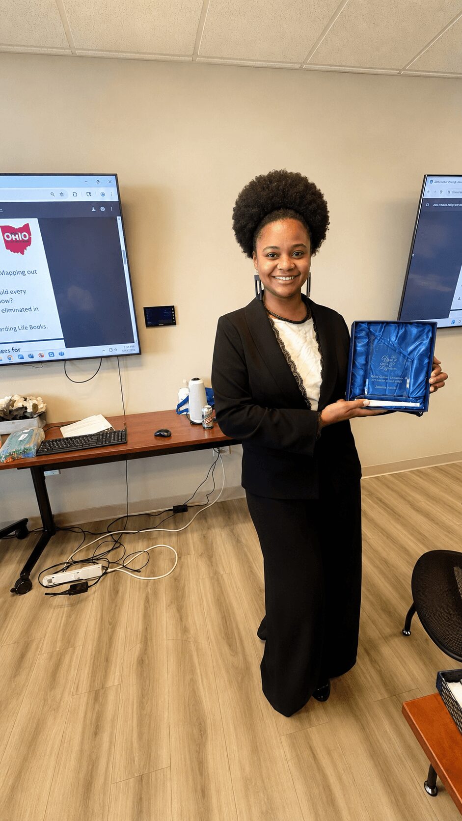 Woman in black suit holding a tablet, smiling, in a conference room with screens and wooden floor.