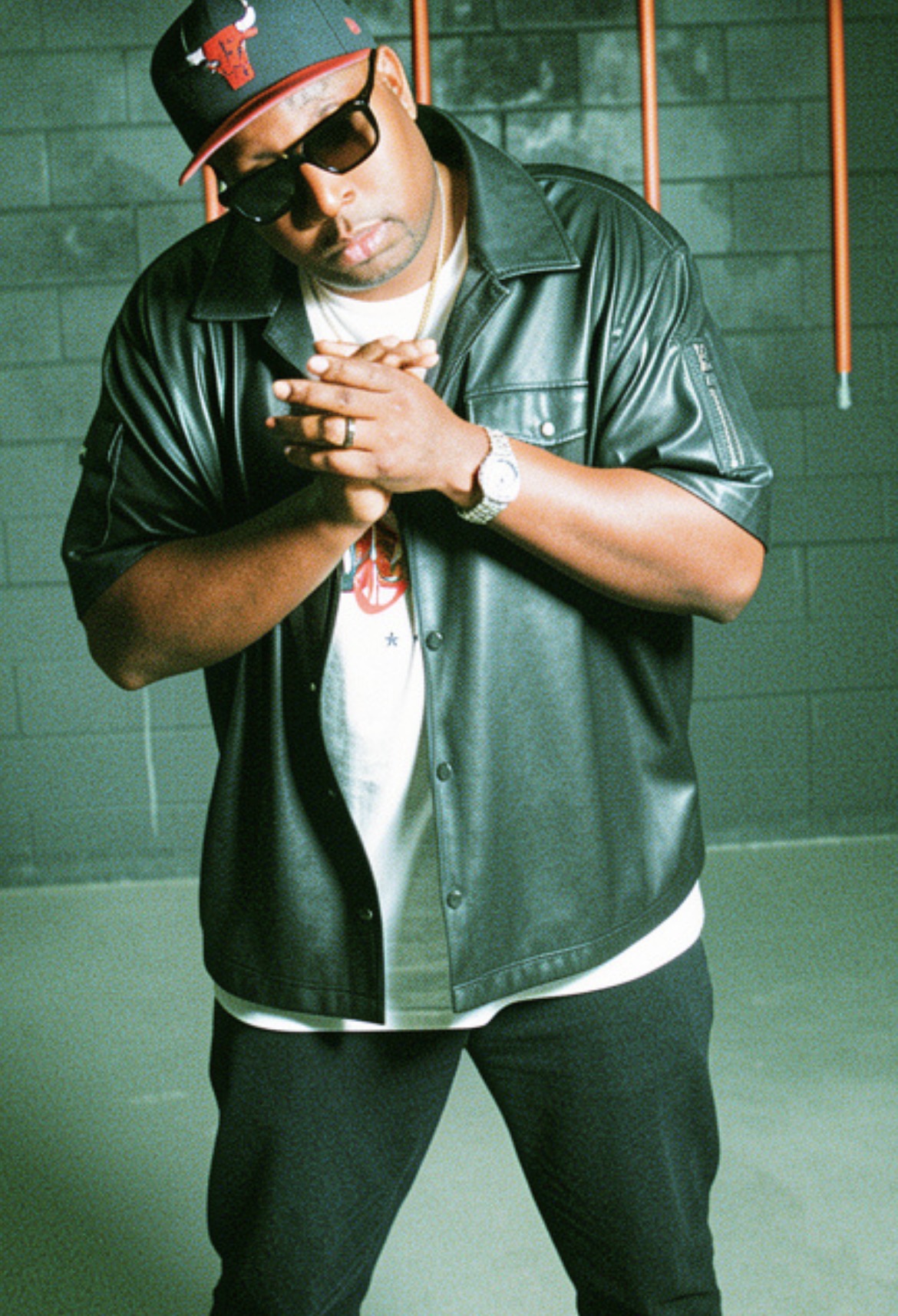Man wearing sunglasses, a cap, leather jacket, and watch, standing with hands clasped in front, against a green wall with orange bars.