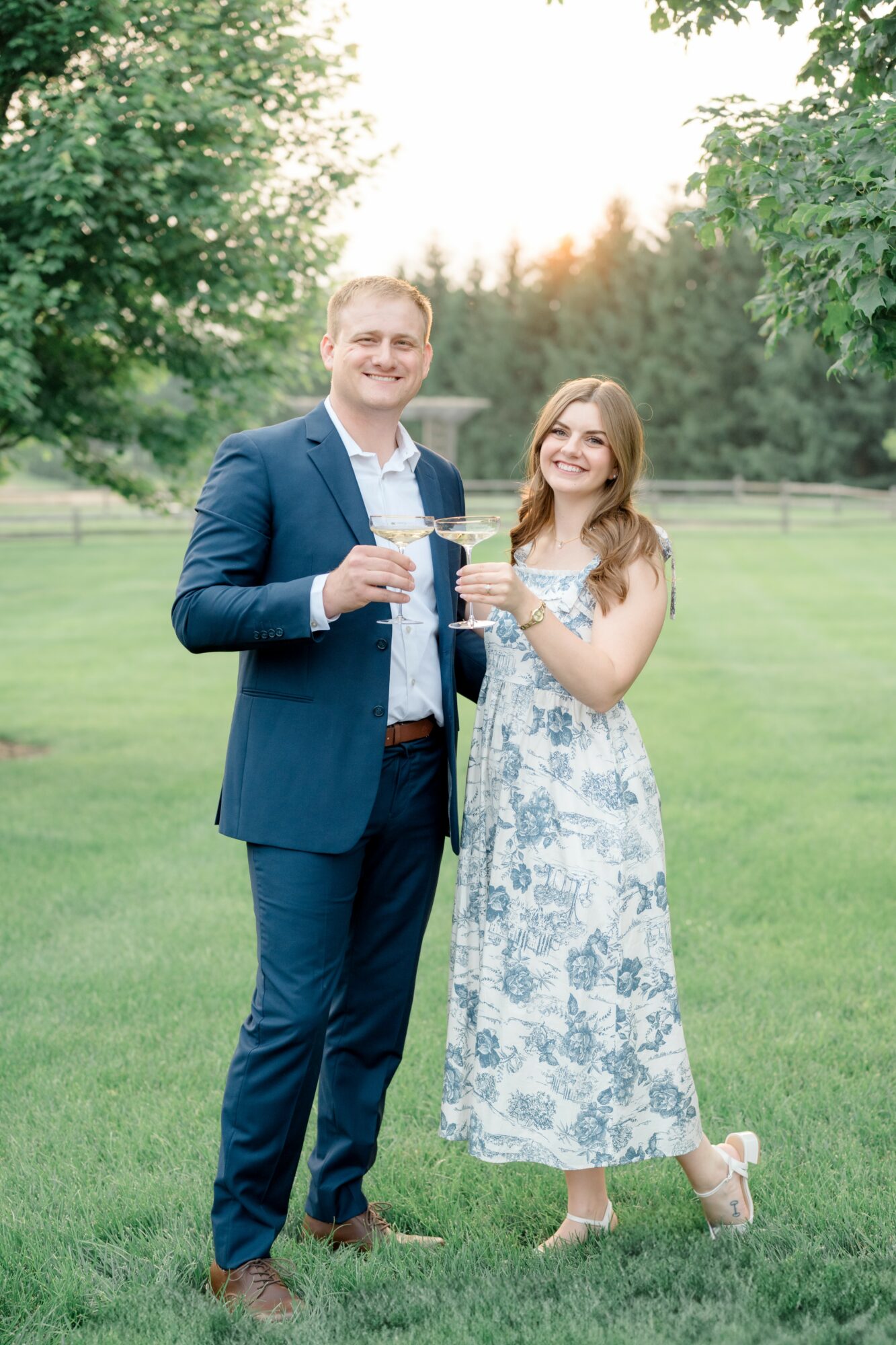 A man and woman in formal attire holding glasses outdoors on a grassy field with trees, smiling at the camera.