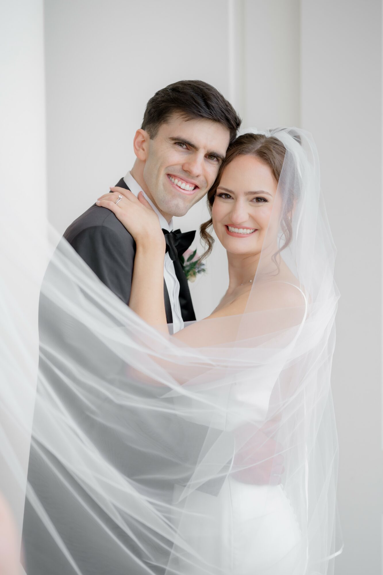 A smiling bride and groom embrace, with the bride wearing a veil and white dress, and the groom in a dark suit.