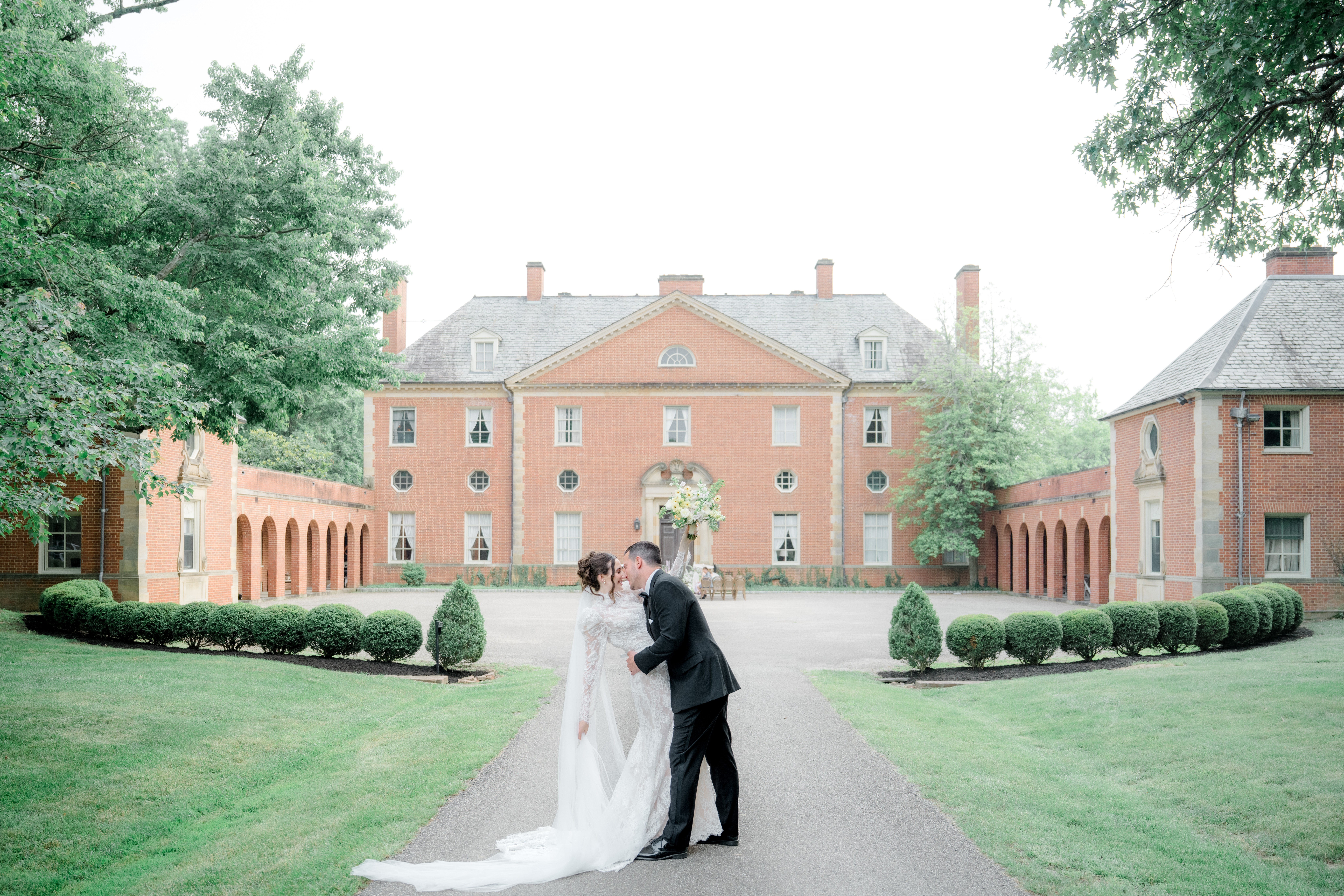 A bride and groom stand on a pathway in front of a large historic building, surrounded by greenery and trees.