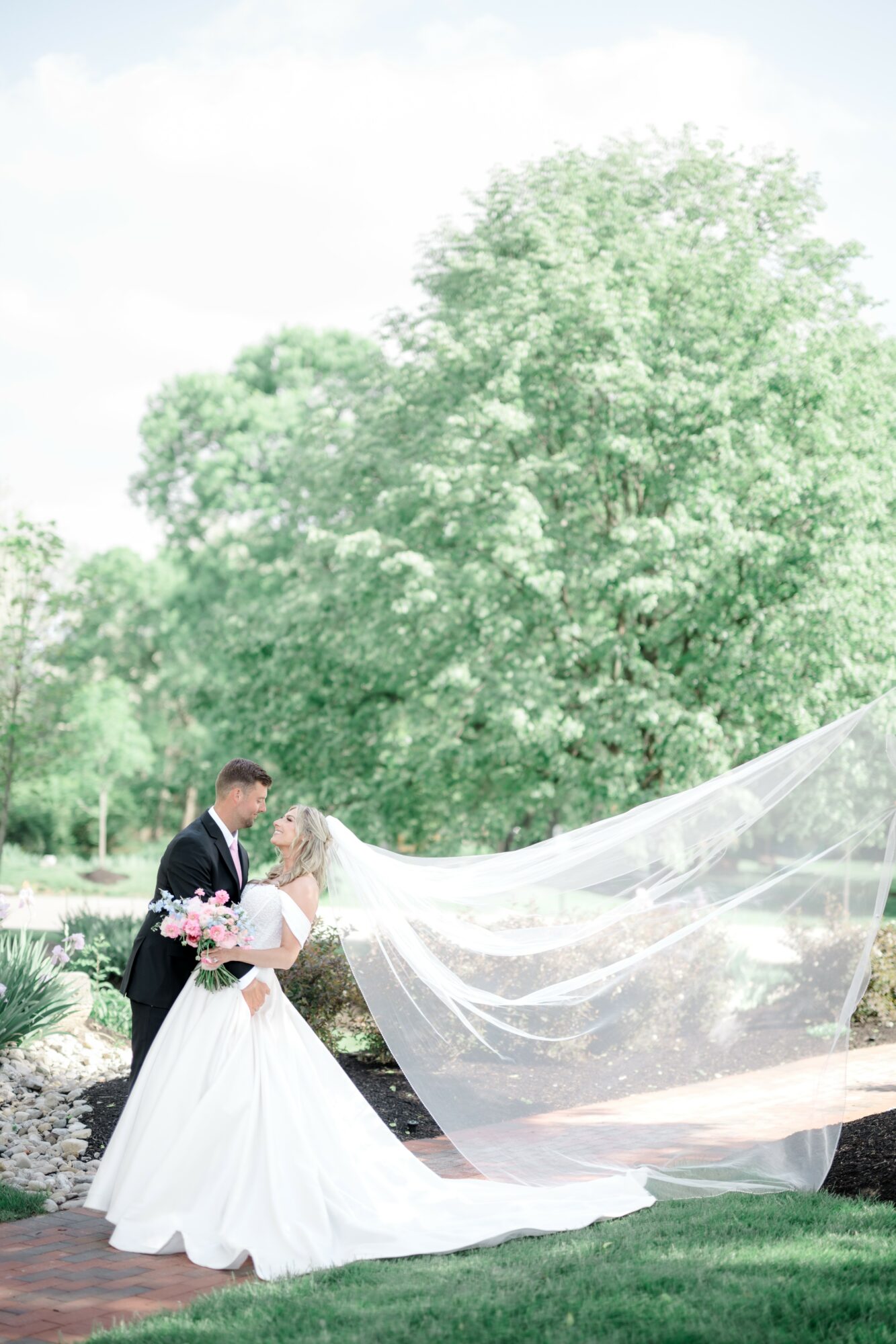 A bride and groom stand outdoors, holding hands, with a large tree and greenery in the background, bride's veil flowing.