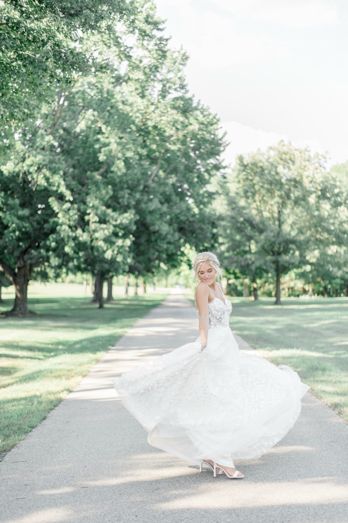 Woman in wedding dress spinning on a park pathway surrounded by trees, sunlight filtering through leaves.