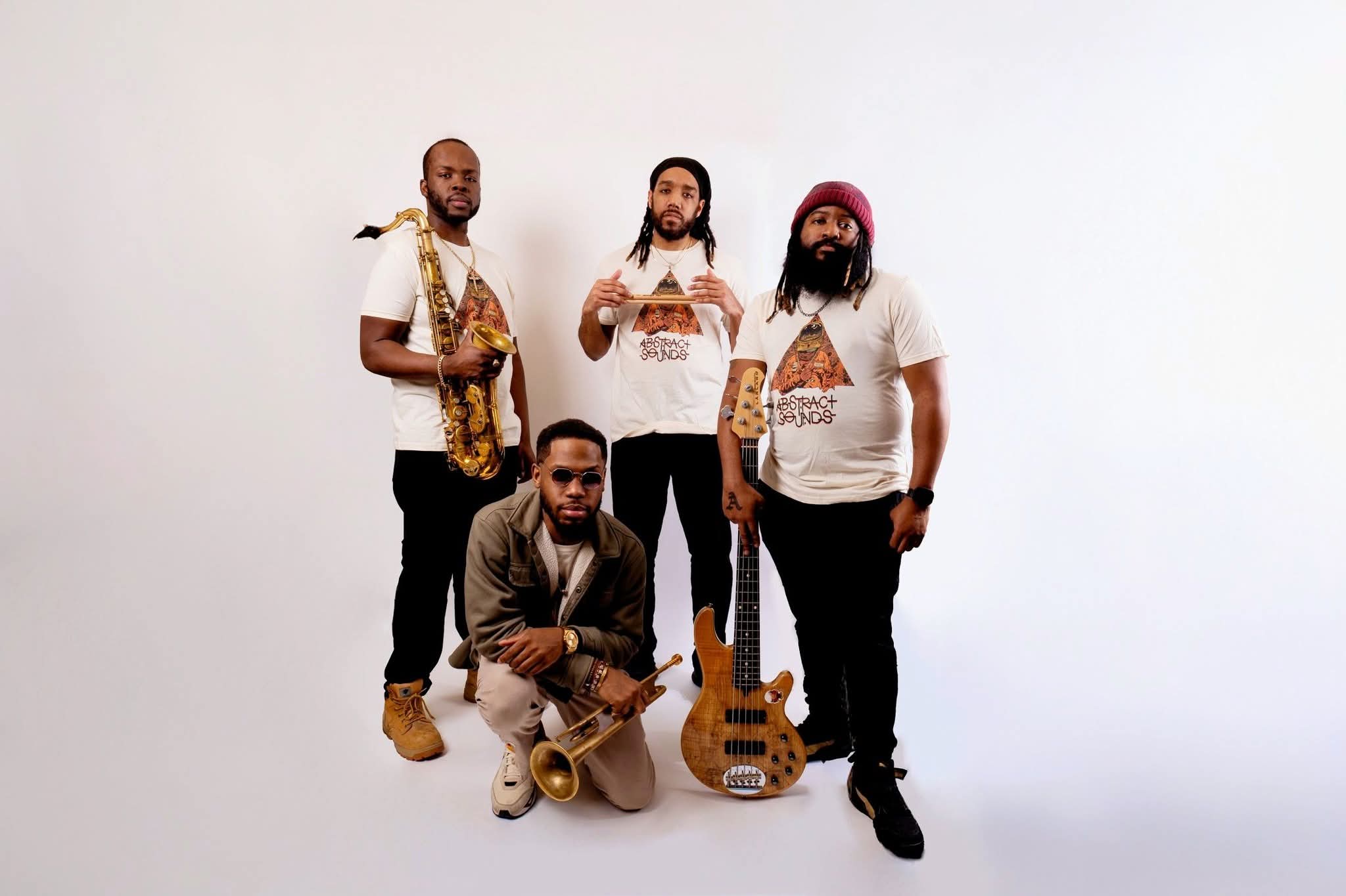Four men pose with musical instruments against a plain white background, one holding a guitar, one with a saxophone, and one with a microphone.