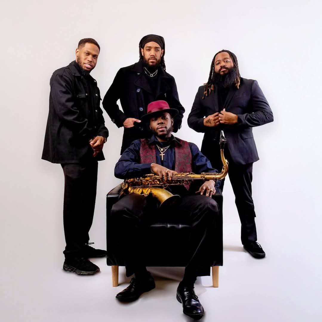Four men pose with one seated holding a saxophone, all dressed in dark clothing, against a plain white background.