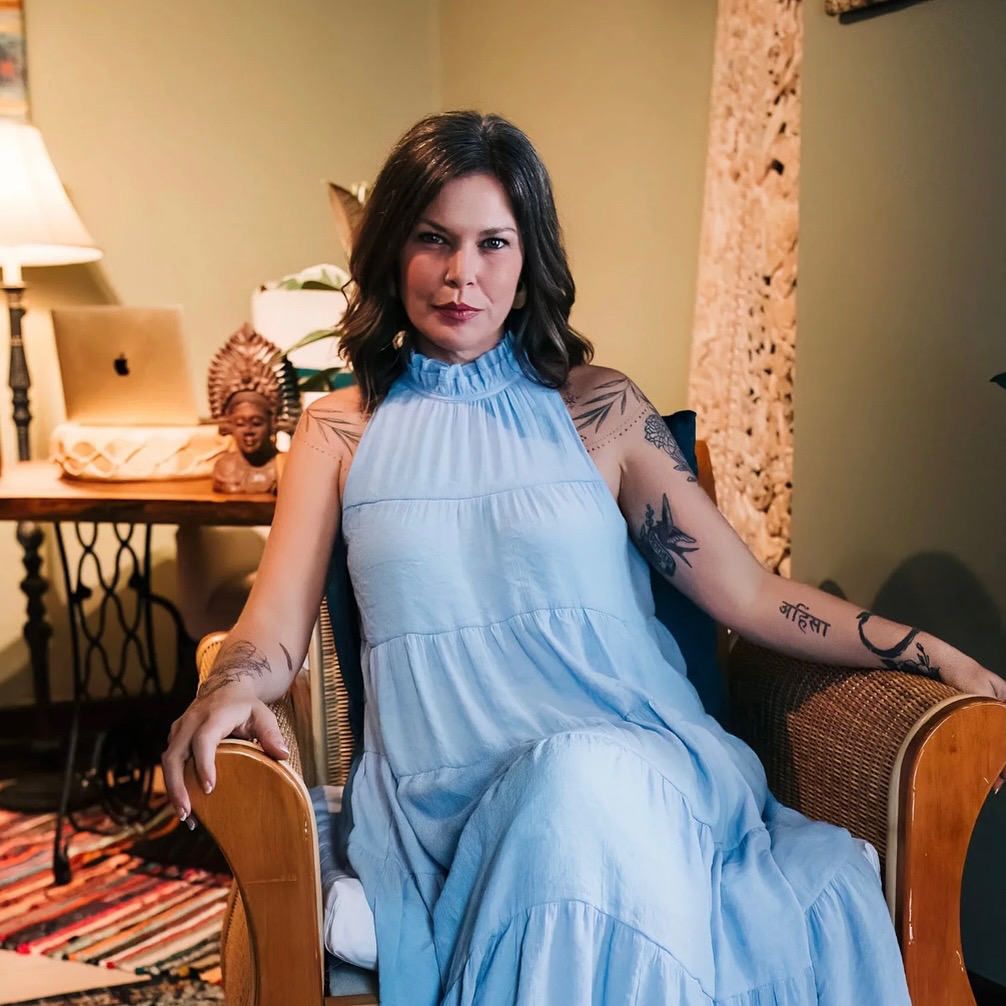 Woman with dark hair in a blue dress sitting in a room with a lamp and curtains, looking at the camera.