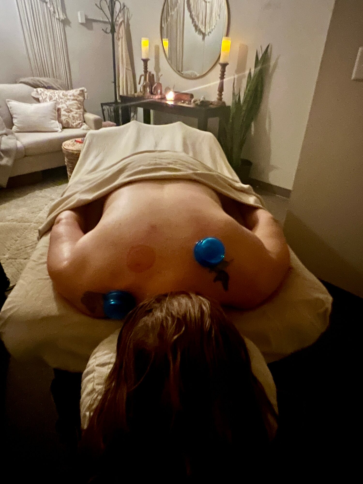 Person lying face down on massage table with blue balls on back, in a cozy room with candles and plants.