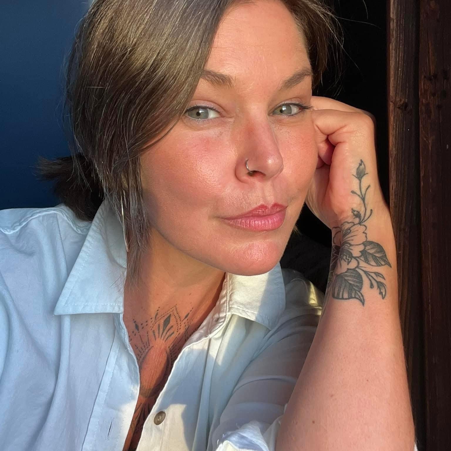 Woman with short brown hair, blue eyes, and a nose piercing, resting her head on her hand, wearing a white shirt.