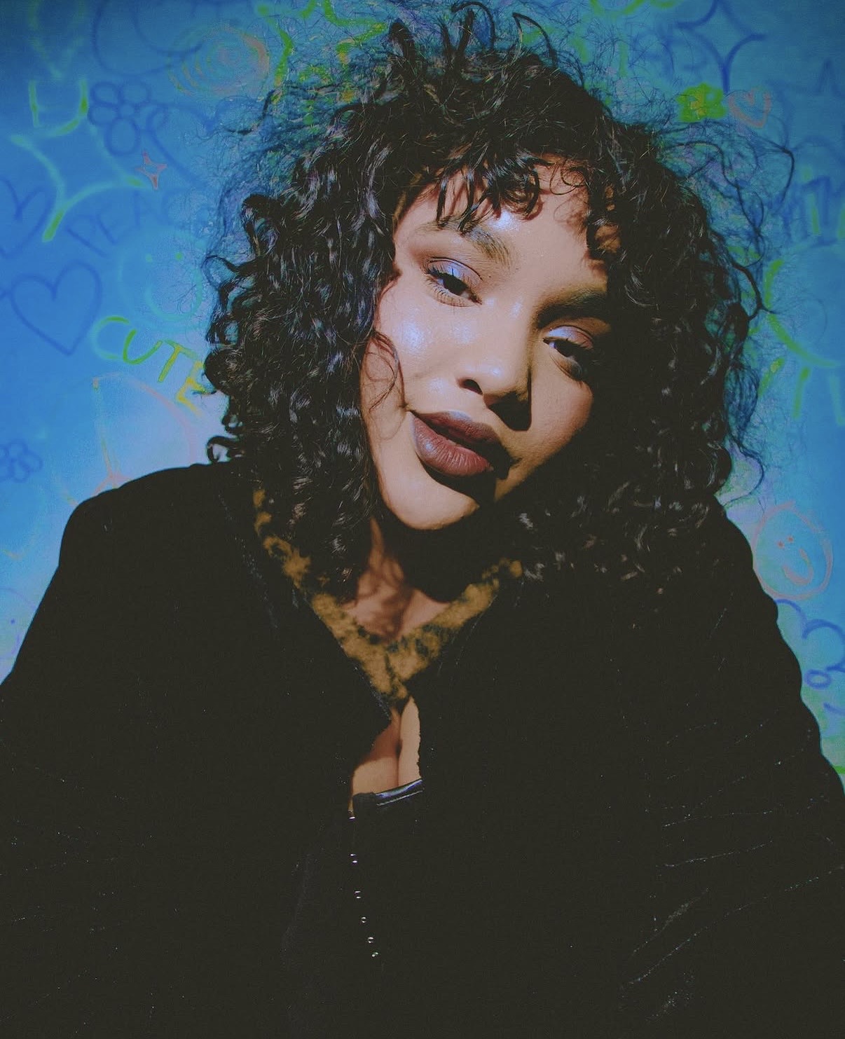 Person with curly hair posing against a colorful background, wearing a dark jacket, looking at the camera.