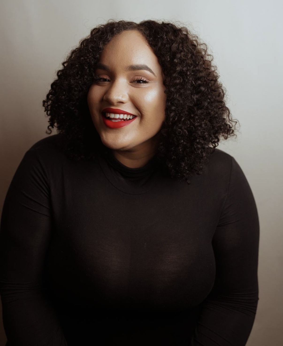 Woman with curly hair smiling, wearing a black top, against a plain background.