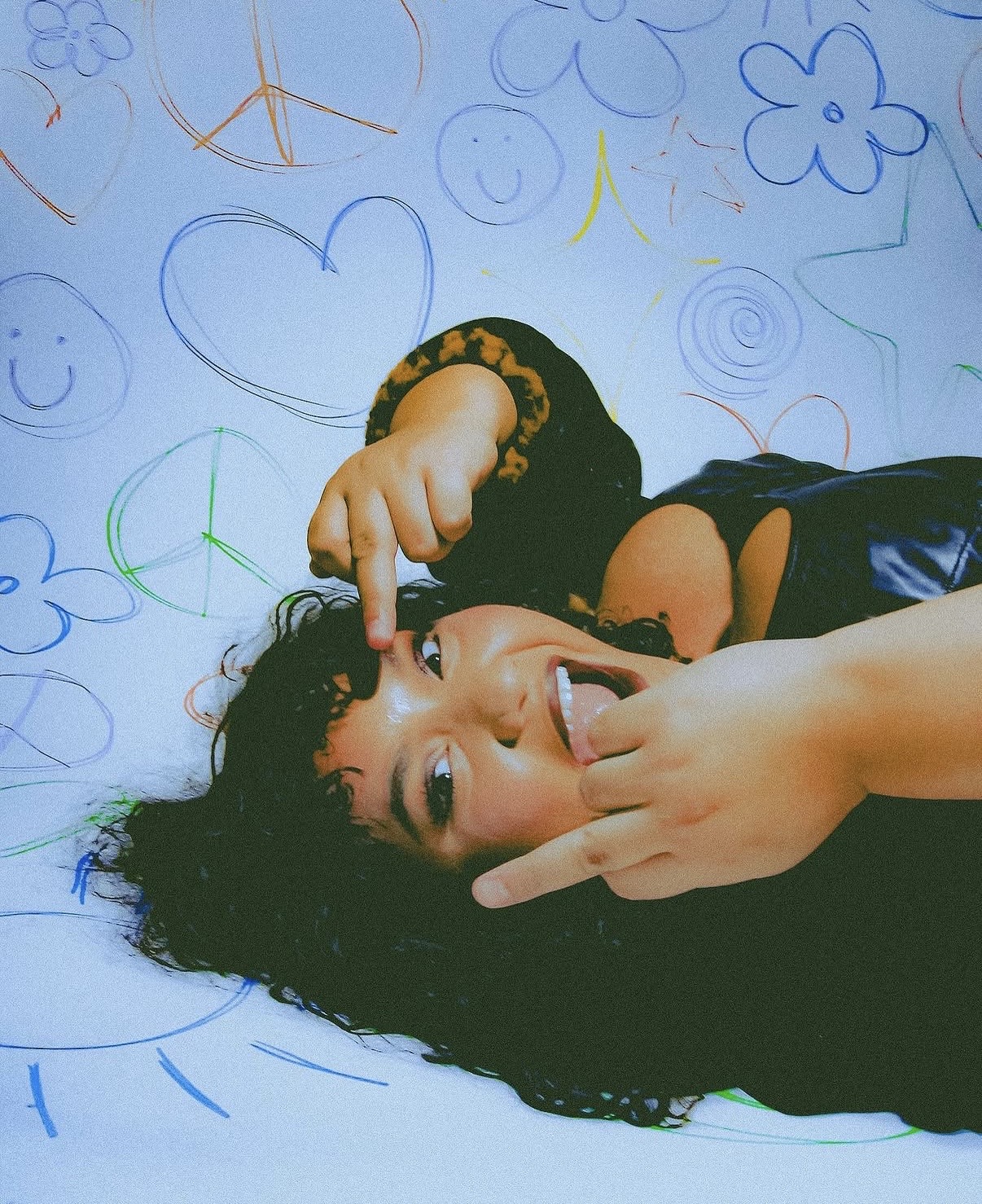 Person lying on the floor with curly hair, smiling, making a peace sign with one hand, surrounded by colorful doodles on the wall.