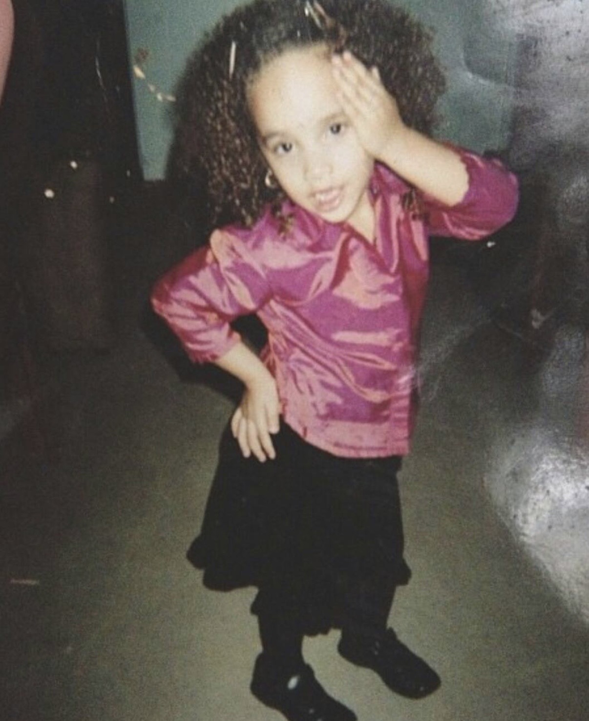 Young girl with curly hair wearing a pink satin top and black skirt, standing with one hand on her head and the other on her hip.