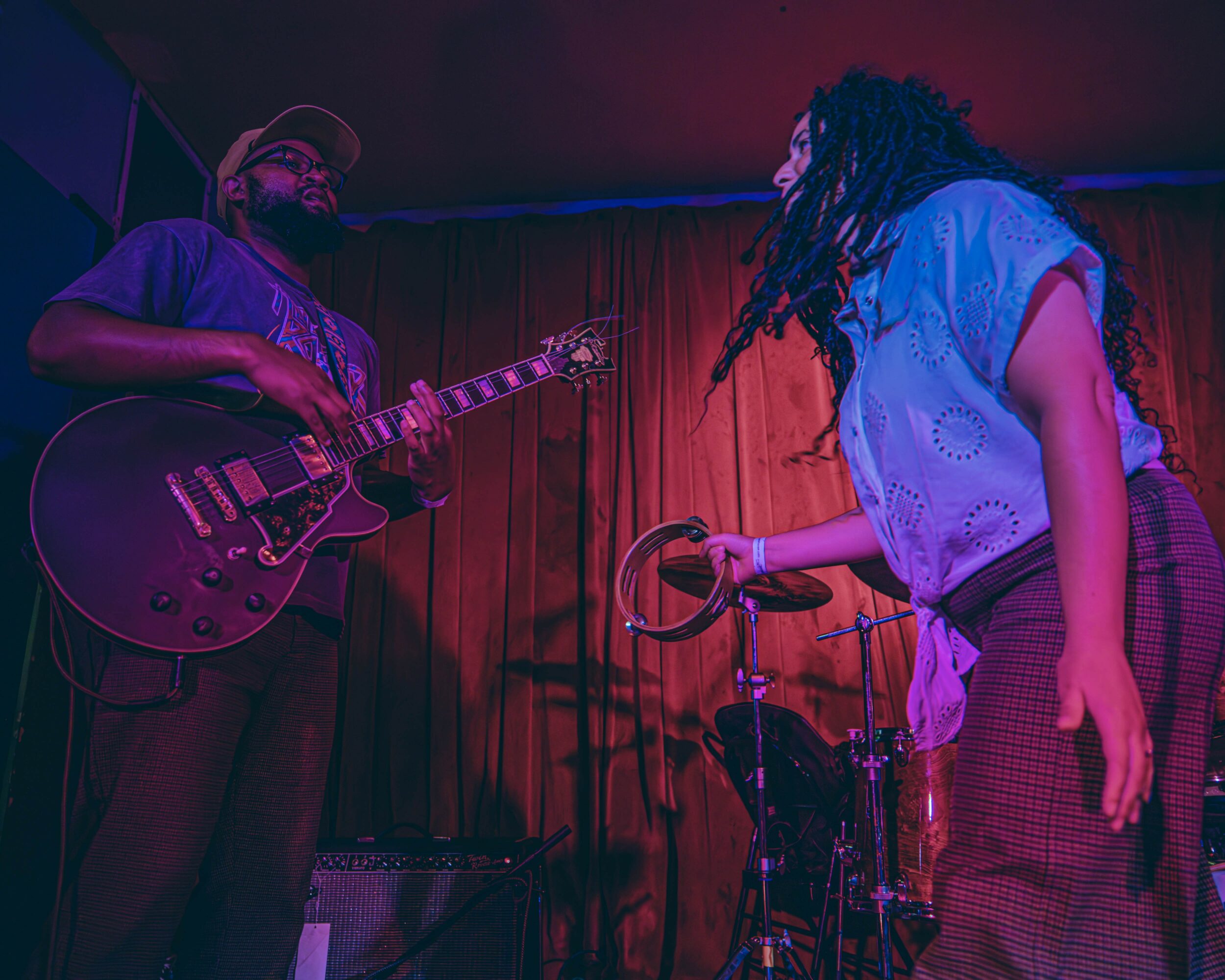 Two women perform on stage with a guitar and tambourine, red curtain background, dim lighting, one with dreadlocks, the other with a hat.