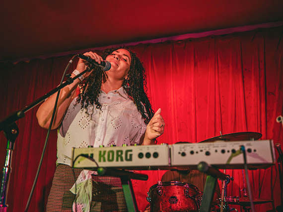 Female singer with curly hair performs on stage with red curtains, a keyboard, and a drum set.