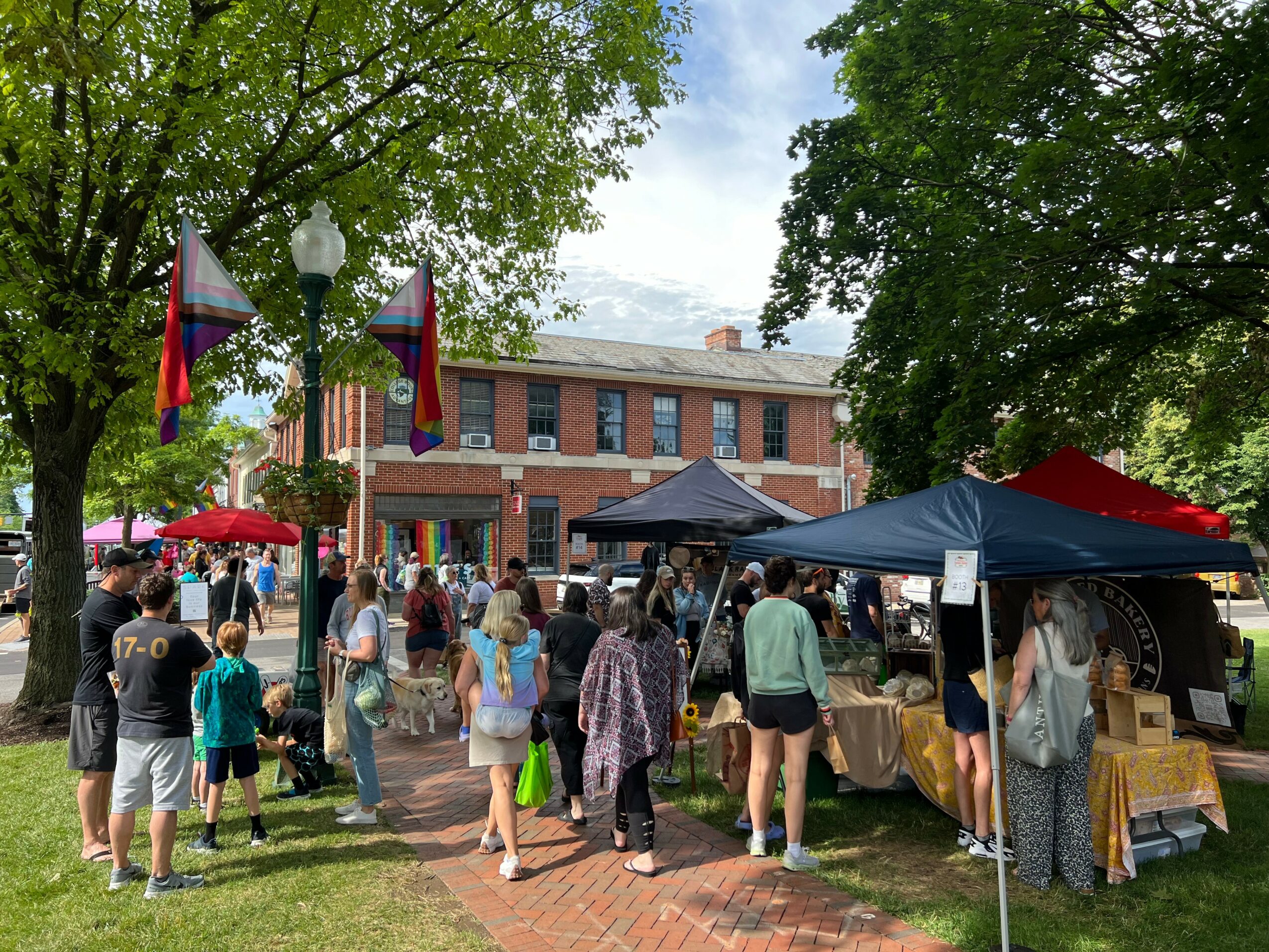 People gather at an outdoor market with tents and tables in front of a brick building, surrounded by trees.