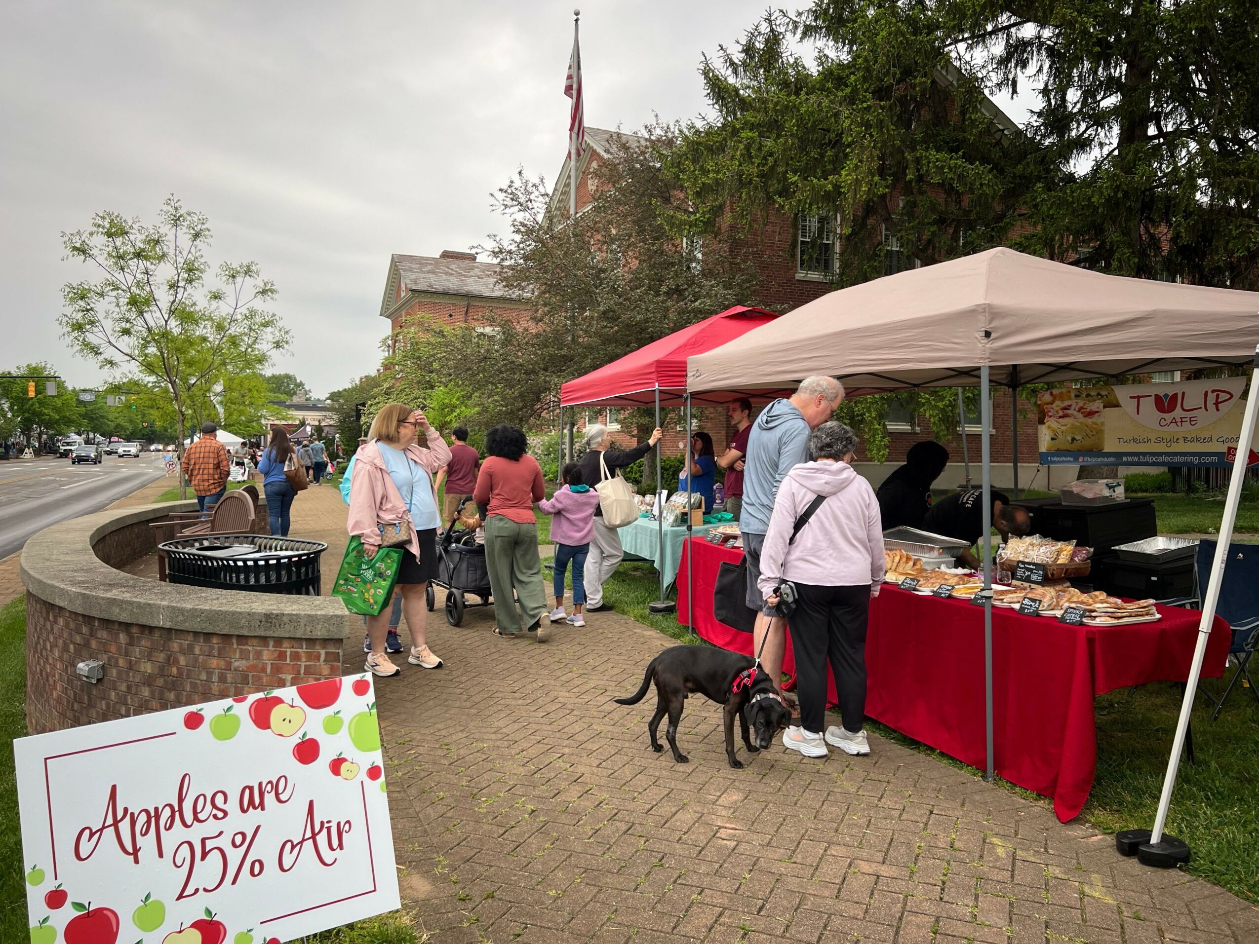 People browsing at an outdoor market with tables and a sign, trees in the background, overcast sky.