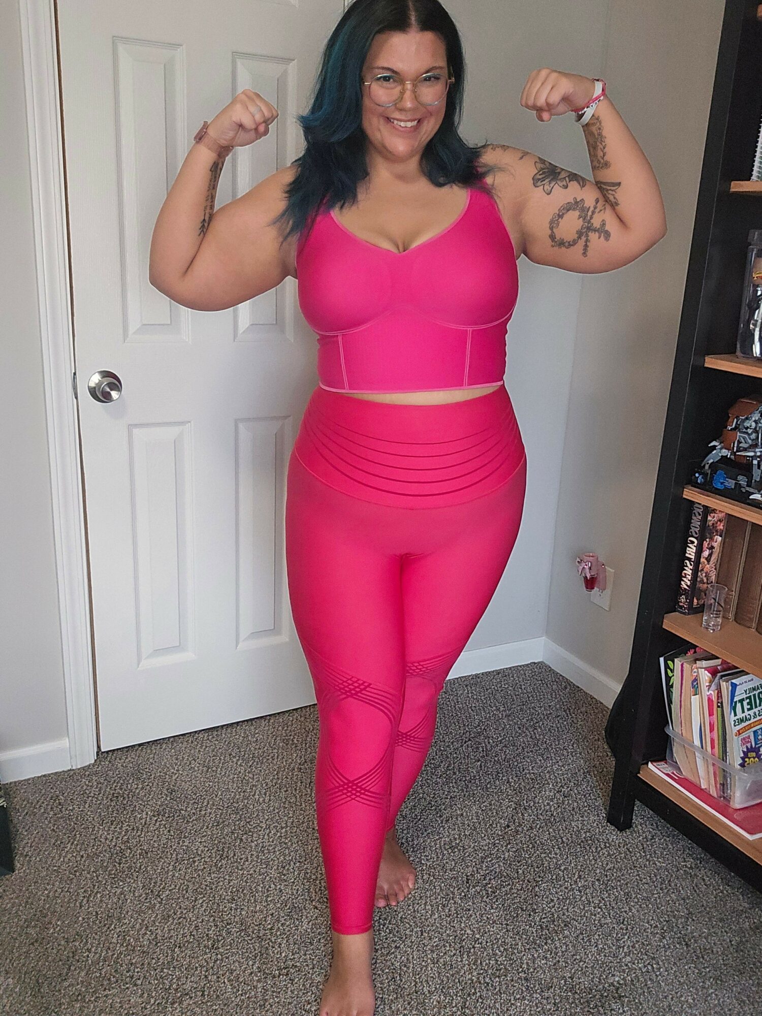 Woman in pink outfit flexing her arms, standing in front of a door with a bookshelf behind her.
