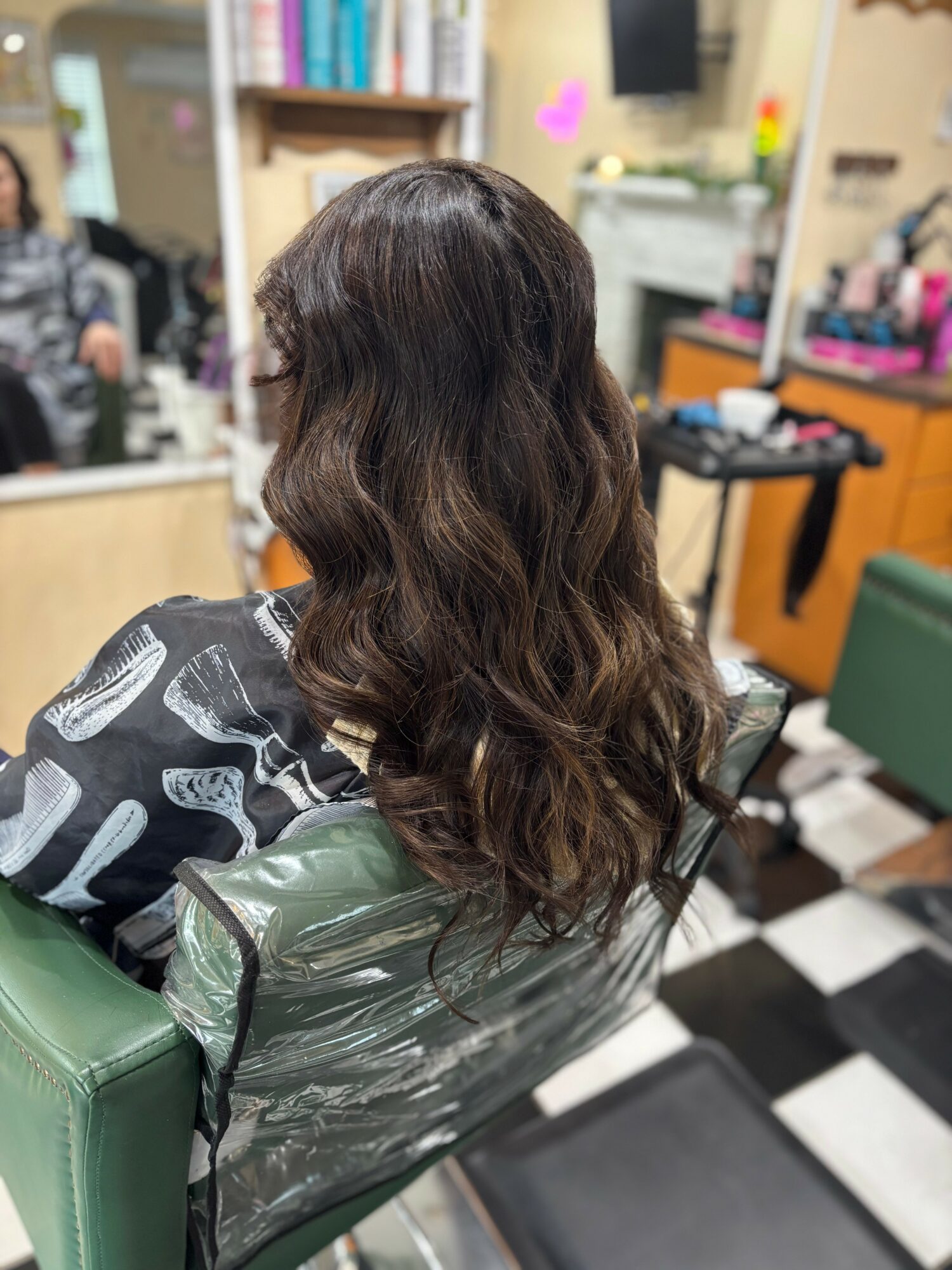 Back of a woman with long, wavy brown hair sitting in a salon chair, surrounded by salon equipment and mirrors.
