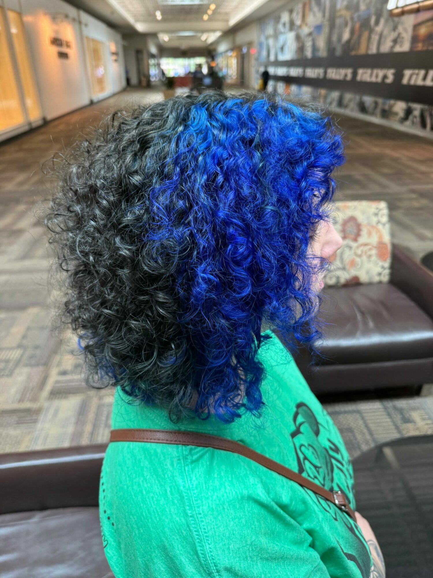 Person with curly hair dyed blue and black, wearing a green shirt, in a shopping mall or store.