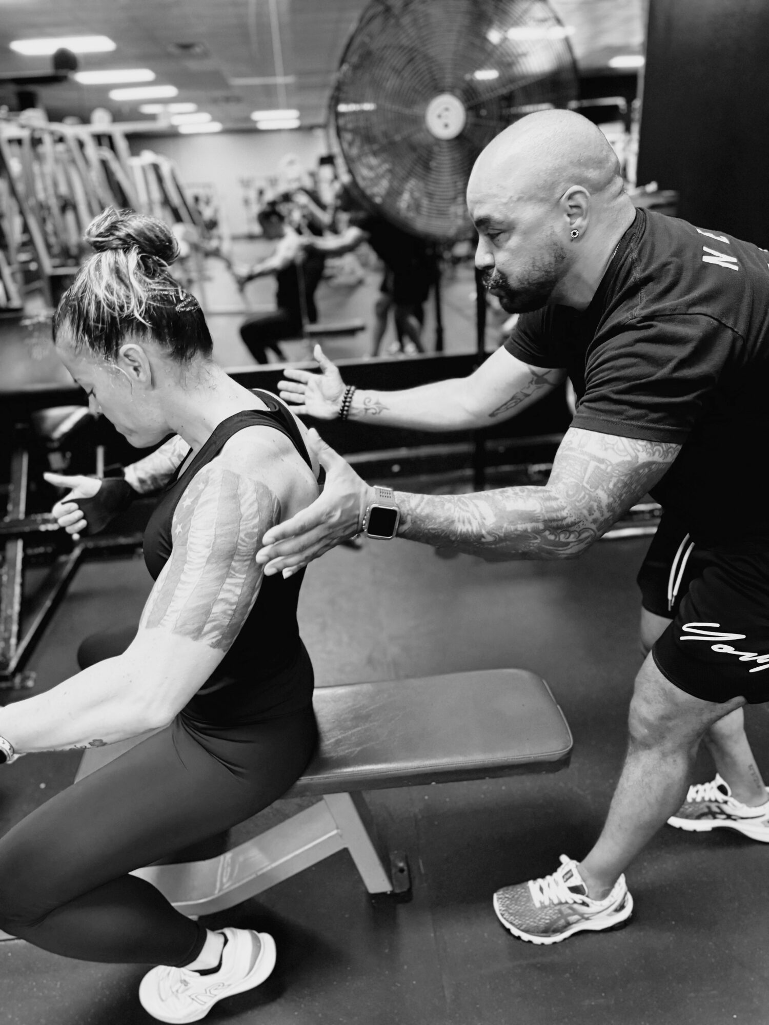 A trainer assists a woman with her workout in a gym, both focused on the exercise, with gym equipment in background.