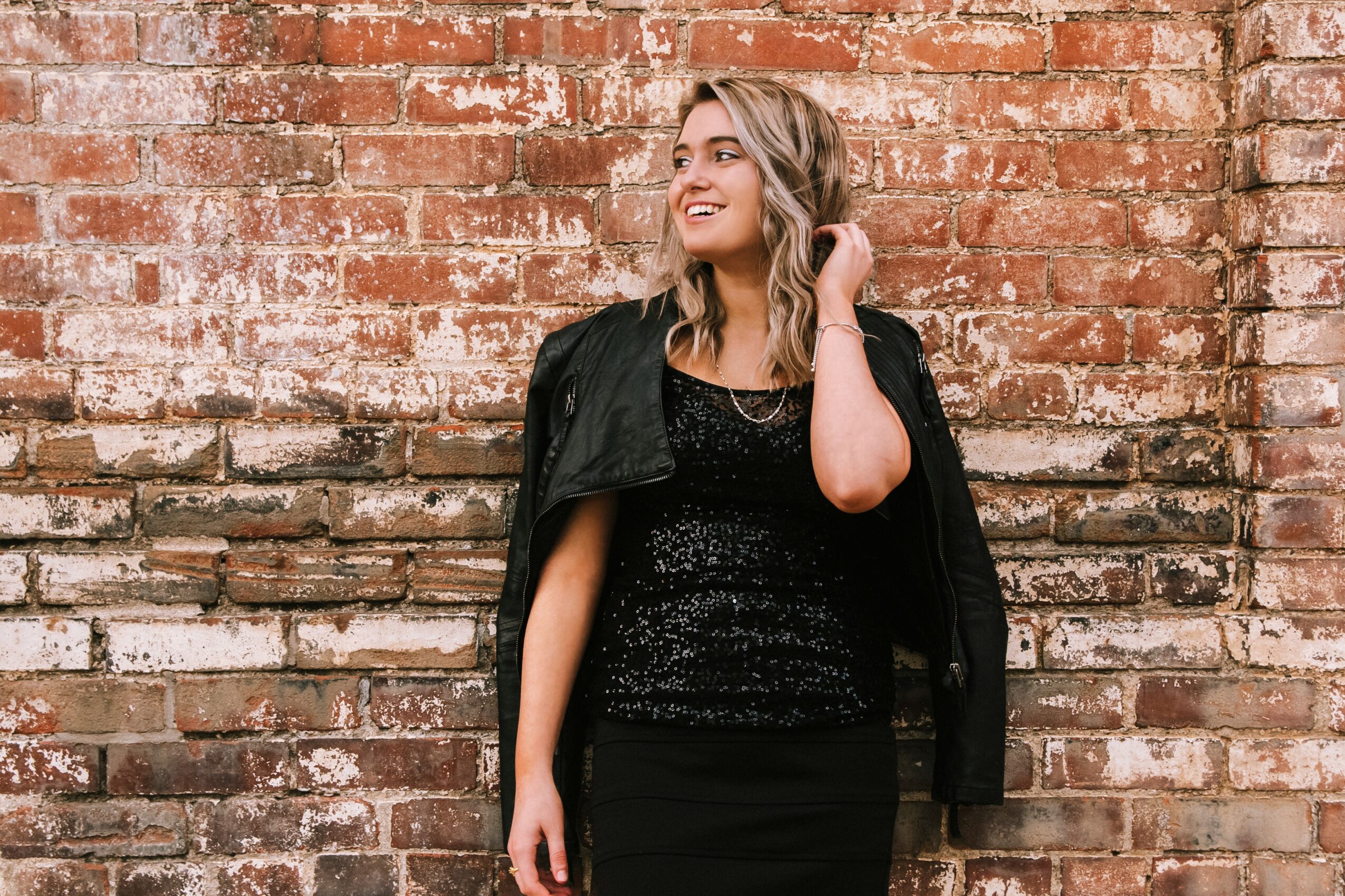 Young woman with blonde hair in a black outfit, standing against a brick wall, smiling and touching her hair.