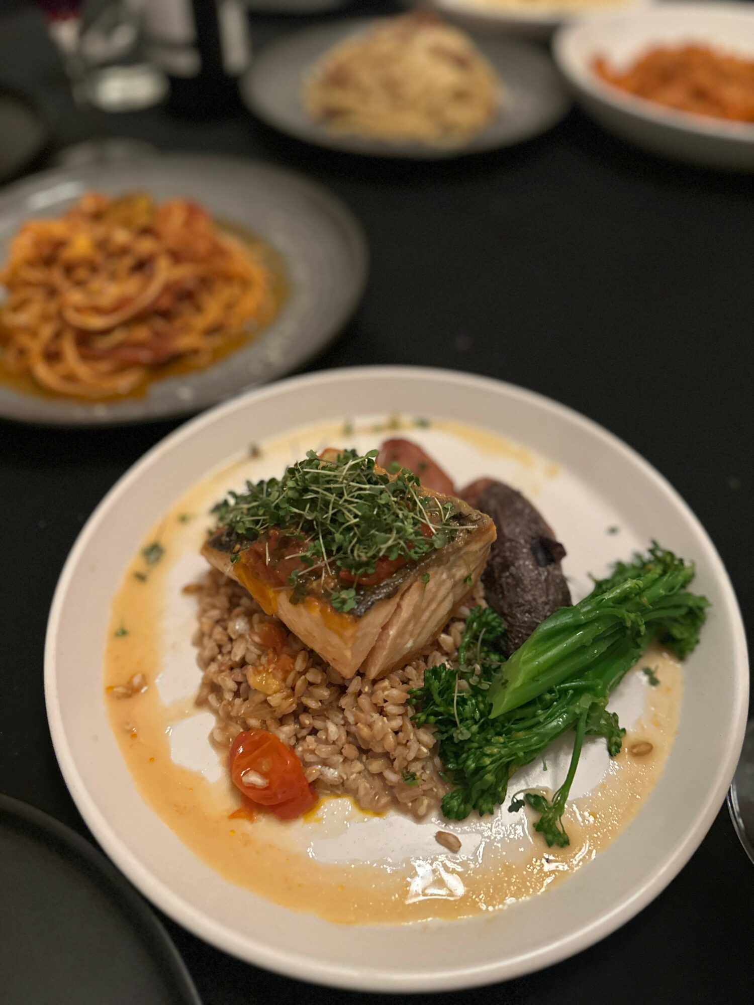 Plate with cooked fish, rice, cherry tomato, greens, and sauce, on a black table with other dishes in background.
