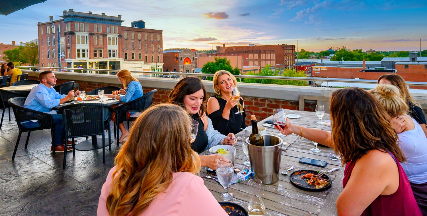 Group of people dining outdoors on a rooftop with city buildings in the background, enjoying food and drinks.