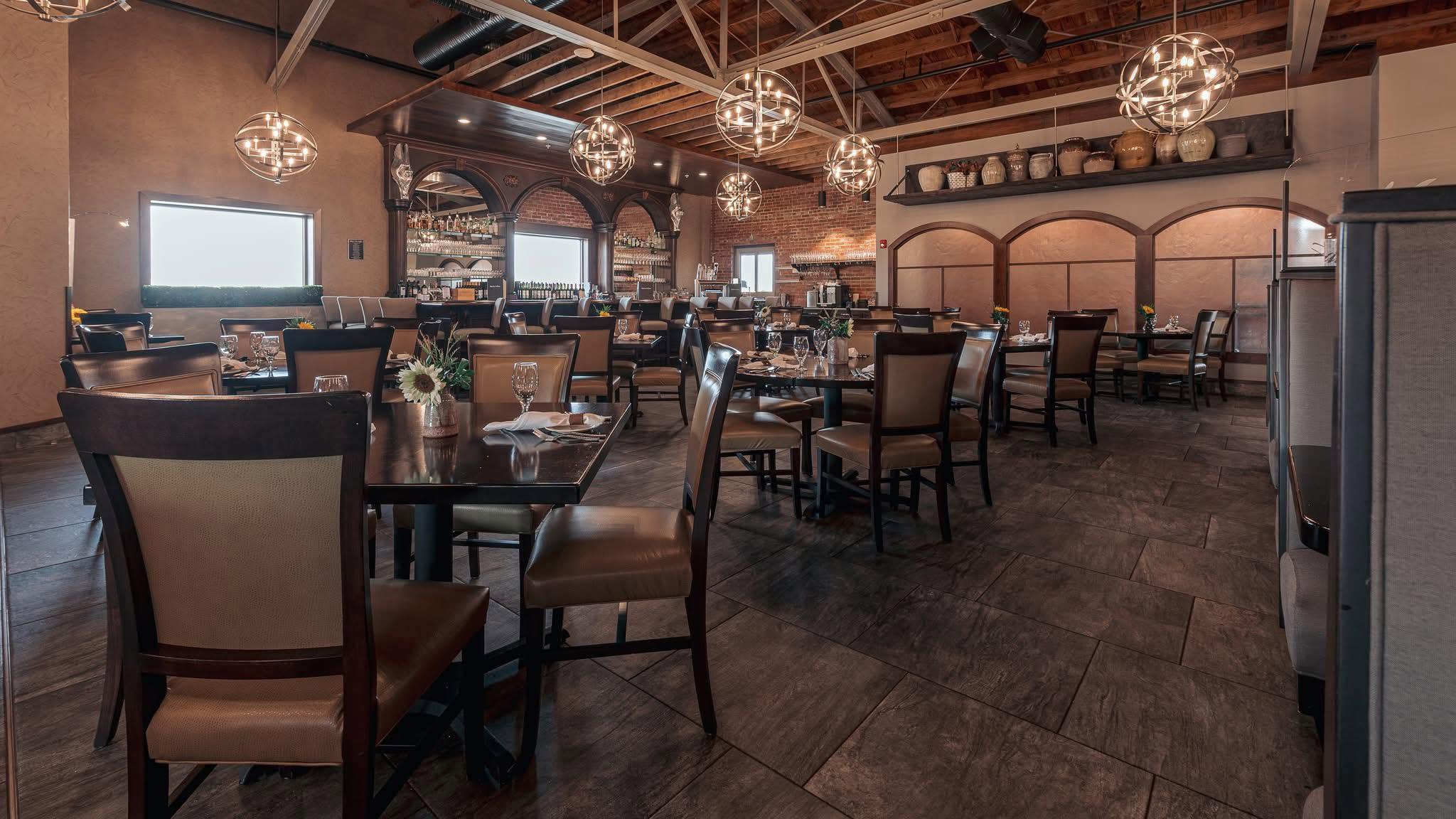 Restaurant interior with tables, chairs, large windows, and decorative lighting fixtures, featuring warm lighting and wooden accents.