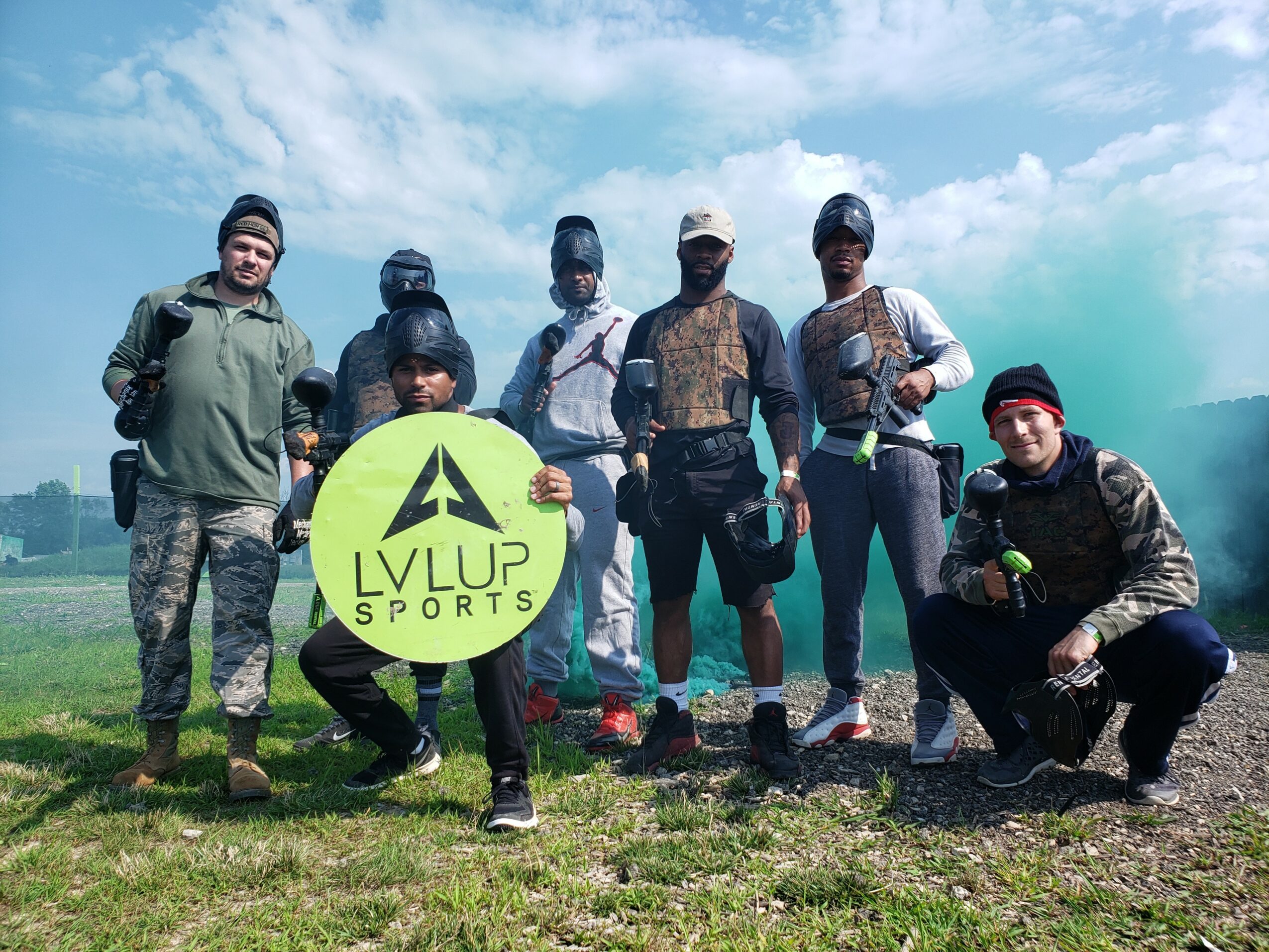 Group of nine people outdoors holding a bright green sign with black text and logo, some wearing paintball gear, on grassy field with blue sky.