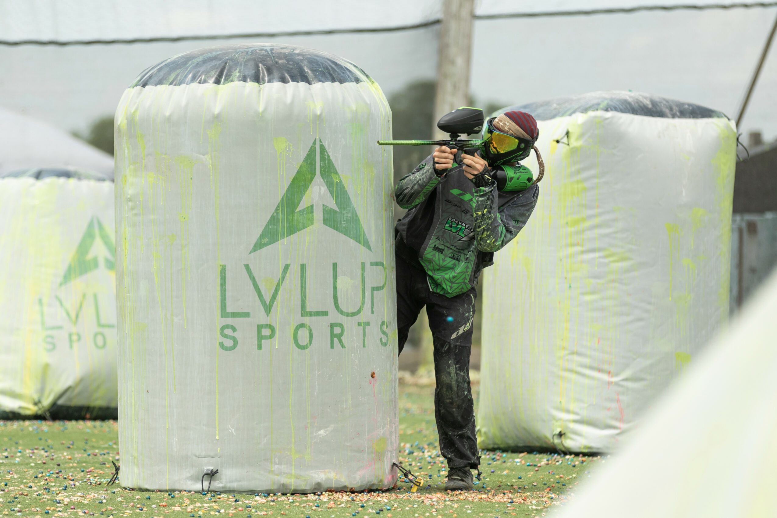 Person wearing a green jacket and black pants aiming a paintball gun behind a large inflatable bunker, outdoor setting.