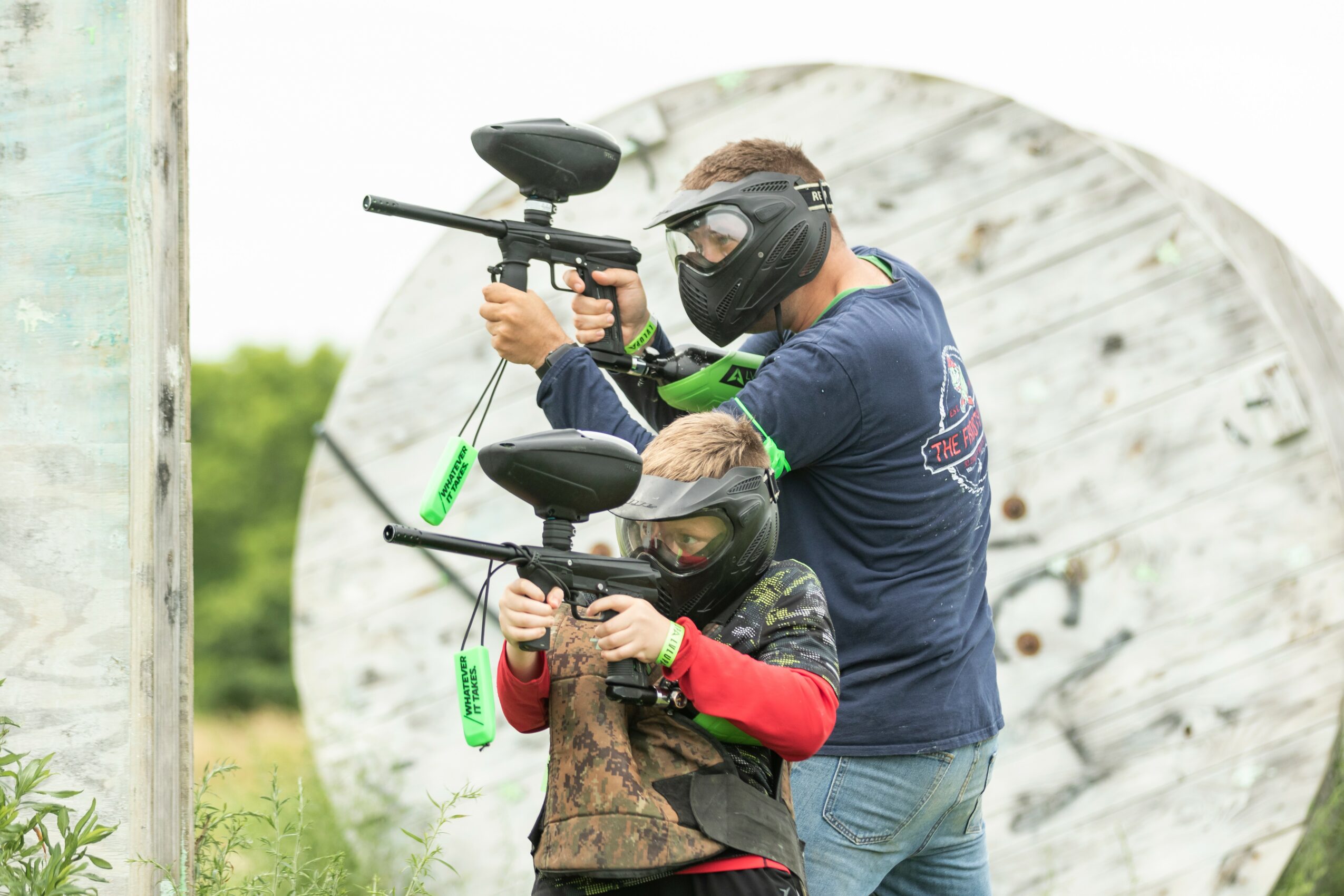 Two people wearing protective masks and goggles hold paintball guns outdoors near a large white circular target.