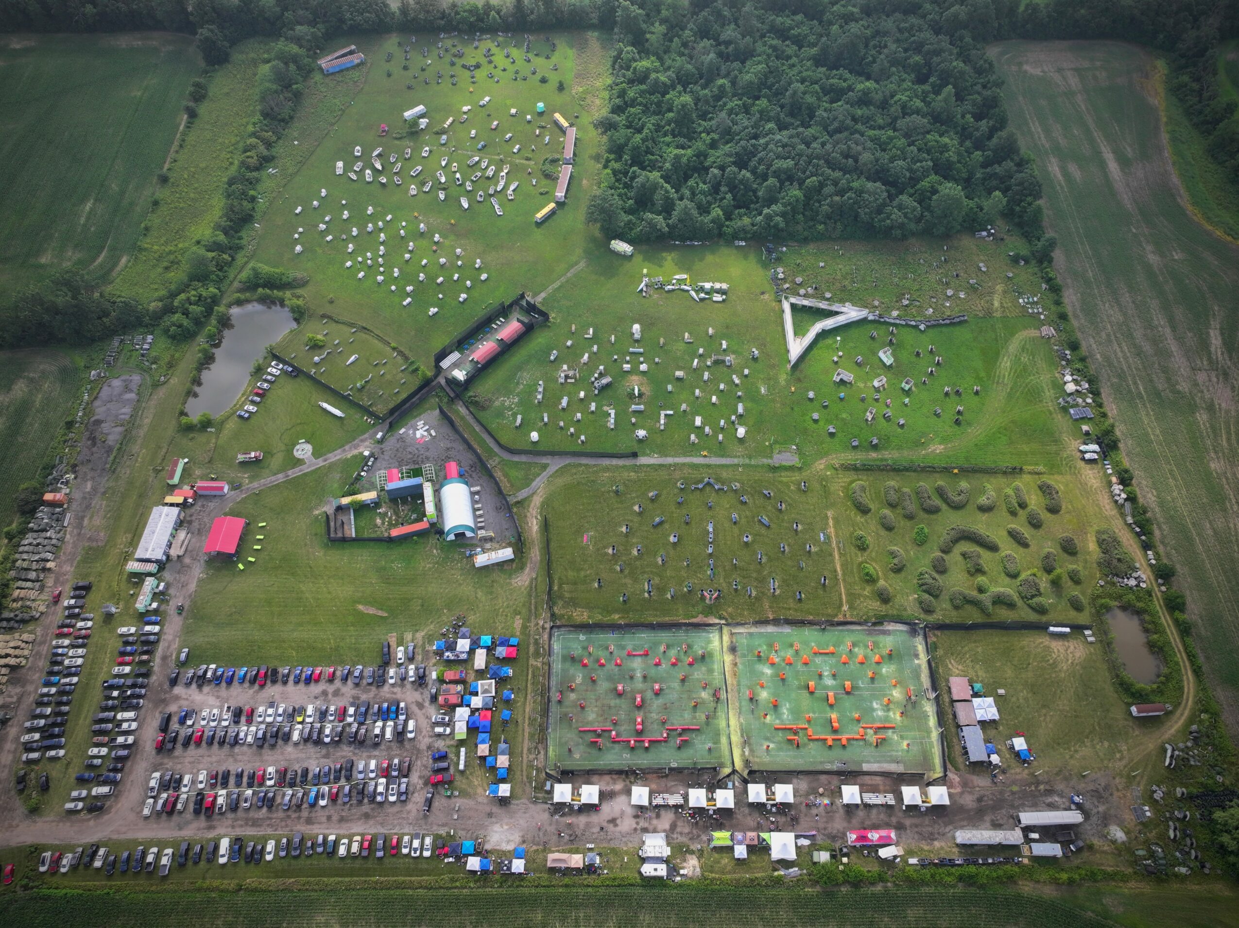 Aerial view of a park with grassy areas, trees, pathways, and a parking lot at the bottom, with various structures and open spaces.