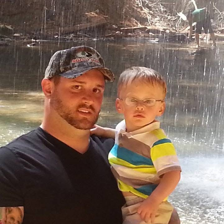Man and young boy standing near a waterfall outdoors, with water falling behind them, in rainy weather.